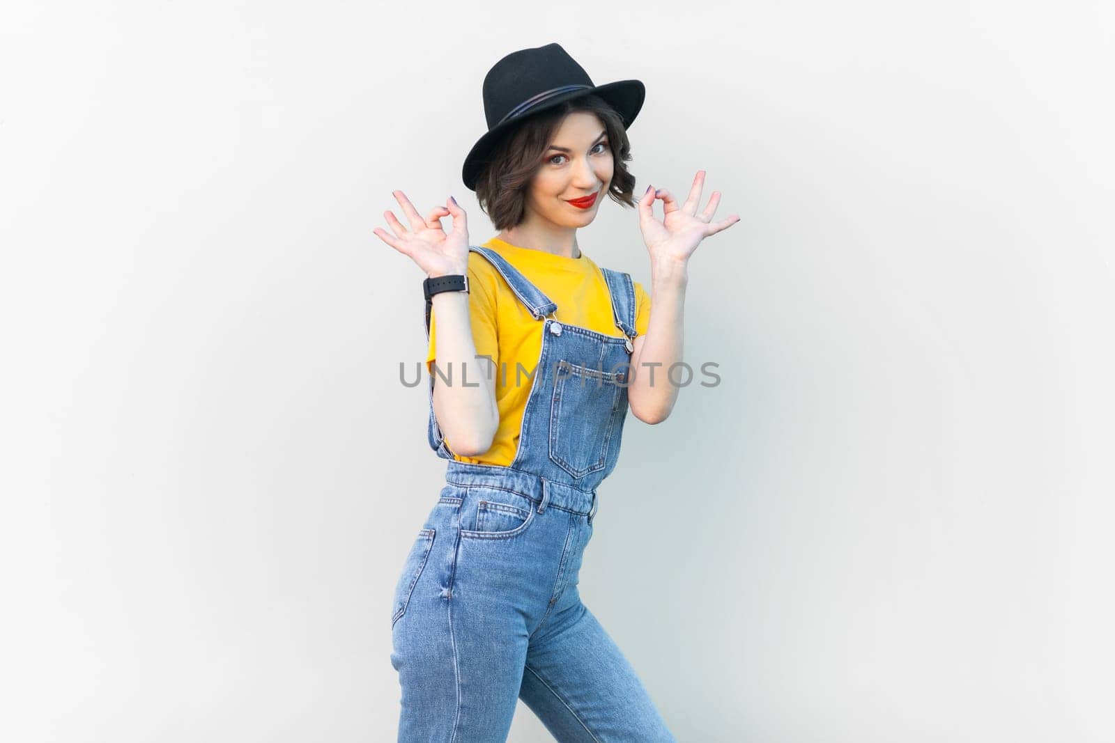 Portrait of optimistic happy hipster woman in blue denim overalls, yellow T-shirt and black hat, raised her arms, showing okay sign. Indoor studio shot isolated on gray background.