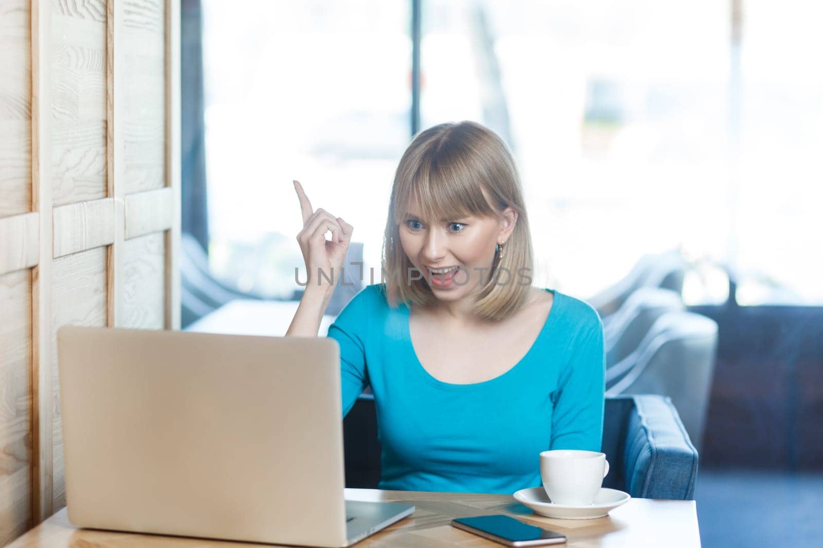 Smart excited positive young woman with blonde hair in blue shirt working on laptop, raised her finger, having idea, knows how to solve problem. Indoor shot in cafe with big window on background.