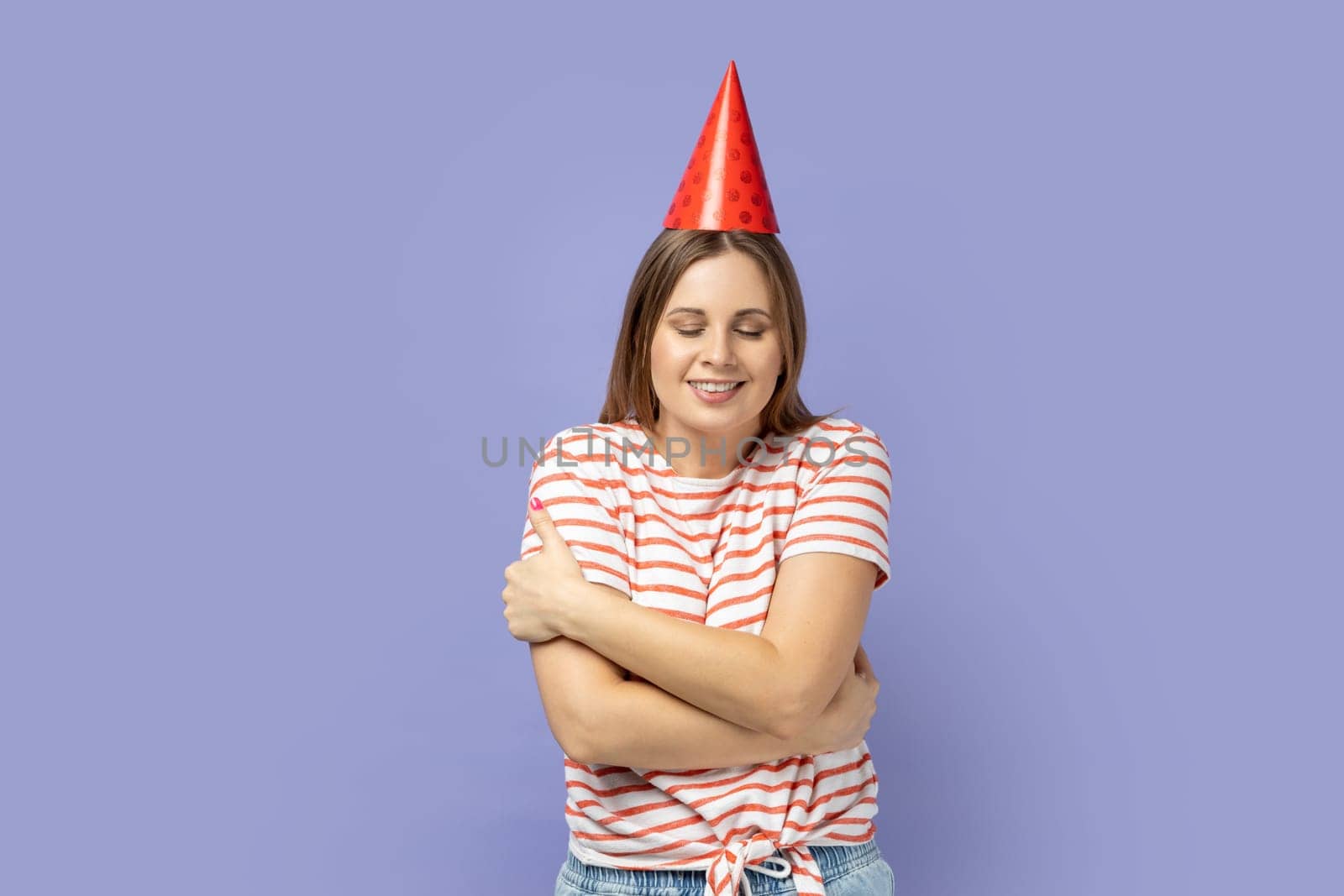 Portrait of self-satisfied egoistic woman in party cone celebrating her birthday, embracing herself and smiling with pleasure, feeling self-pride. Indoor studio shot isolated on purple background.