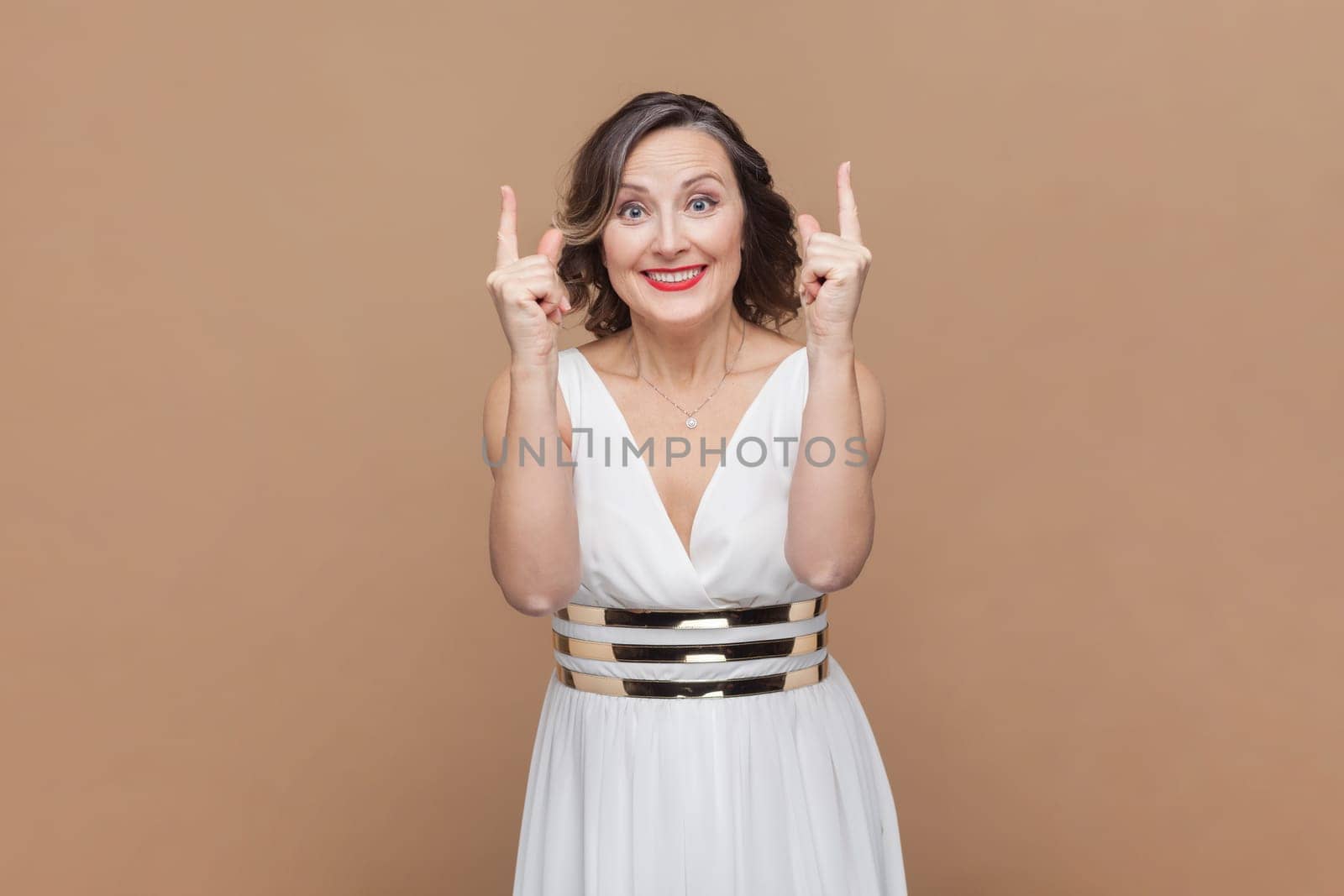 Portrait of smiling happy satisfied middle aged woman with wavy hair pointing upwards and advertisement area, wearing white dress. Indoor studio shot isolated on light brown background.