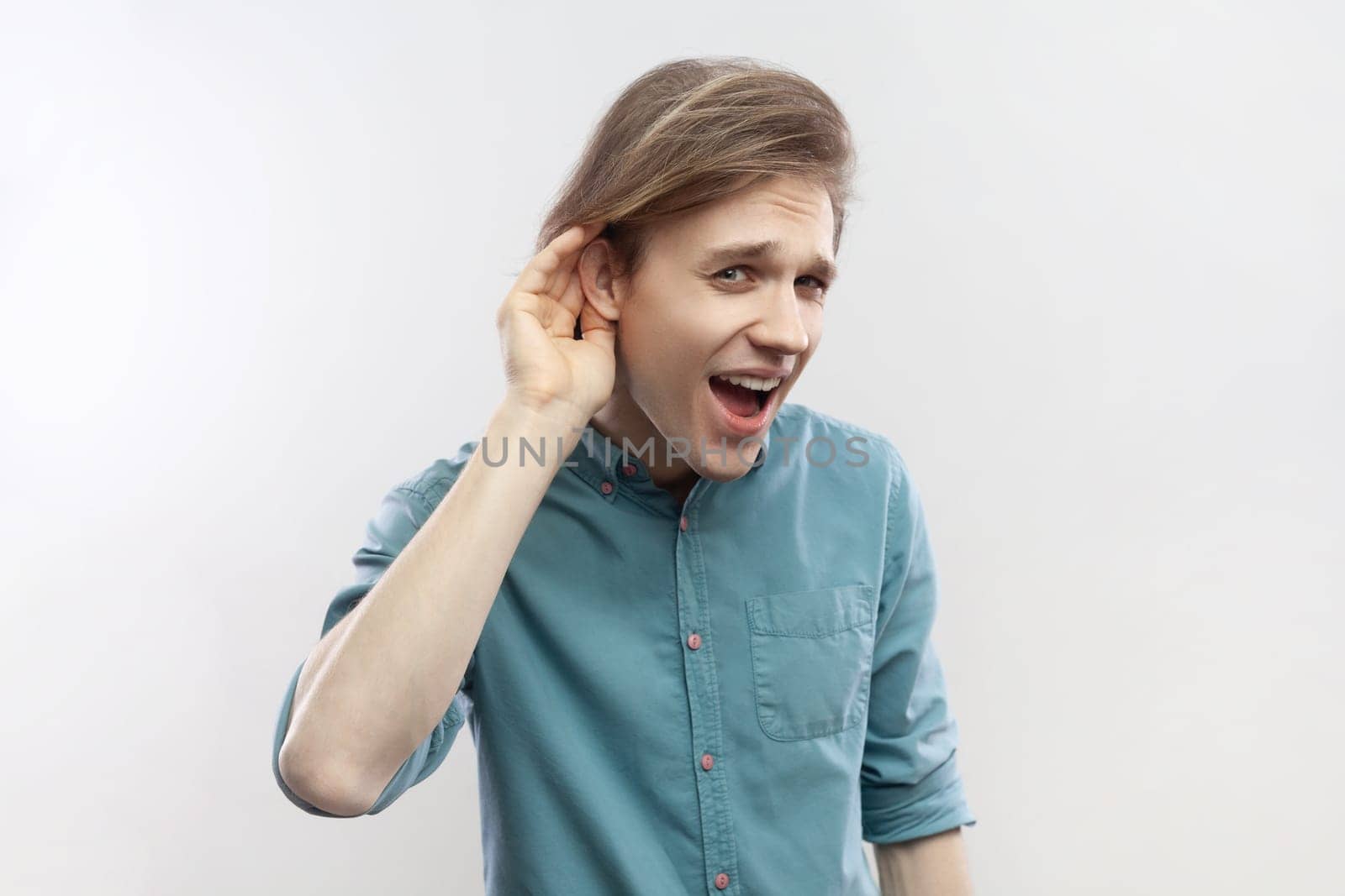 What I dont hear you. Portrait of man standing with hand near ear and listening to whisper, difficult to understand quiet talk, wearing blue shirt. Indoor studio shot isolated on gray background.