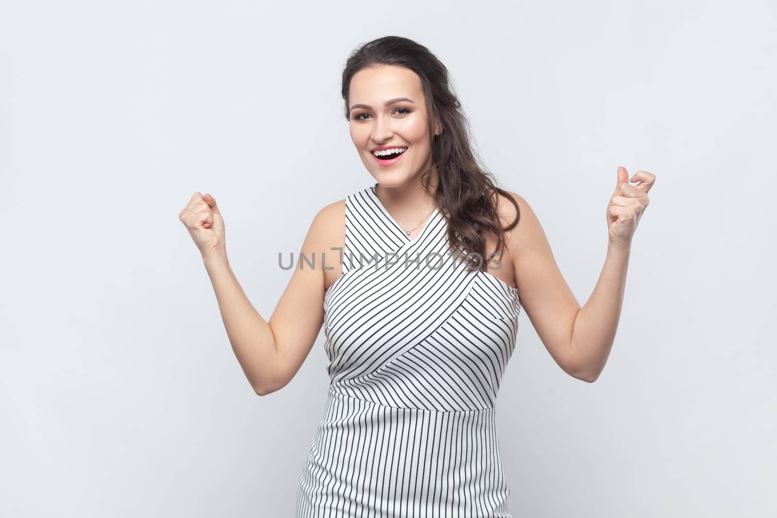 Emotional brunette woman raises clenched fists, exclaims with excitement, rejoices sweet success, feels taste of victory, wearing striped dress. Indoor studio shot isolated on gray background.
