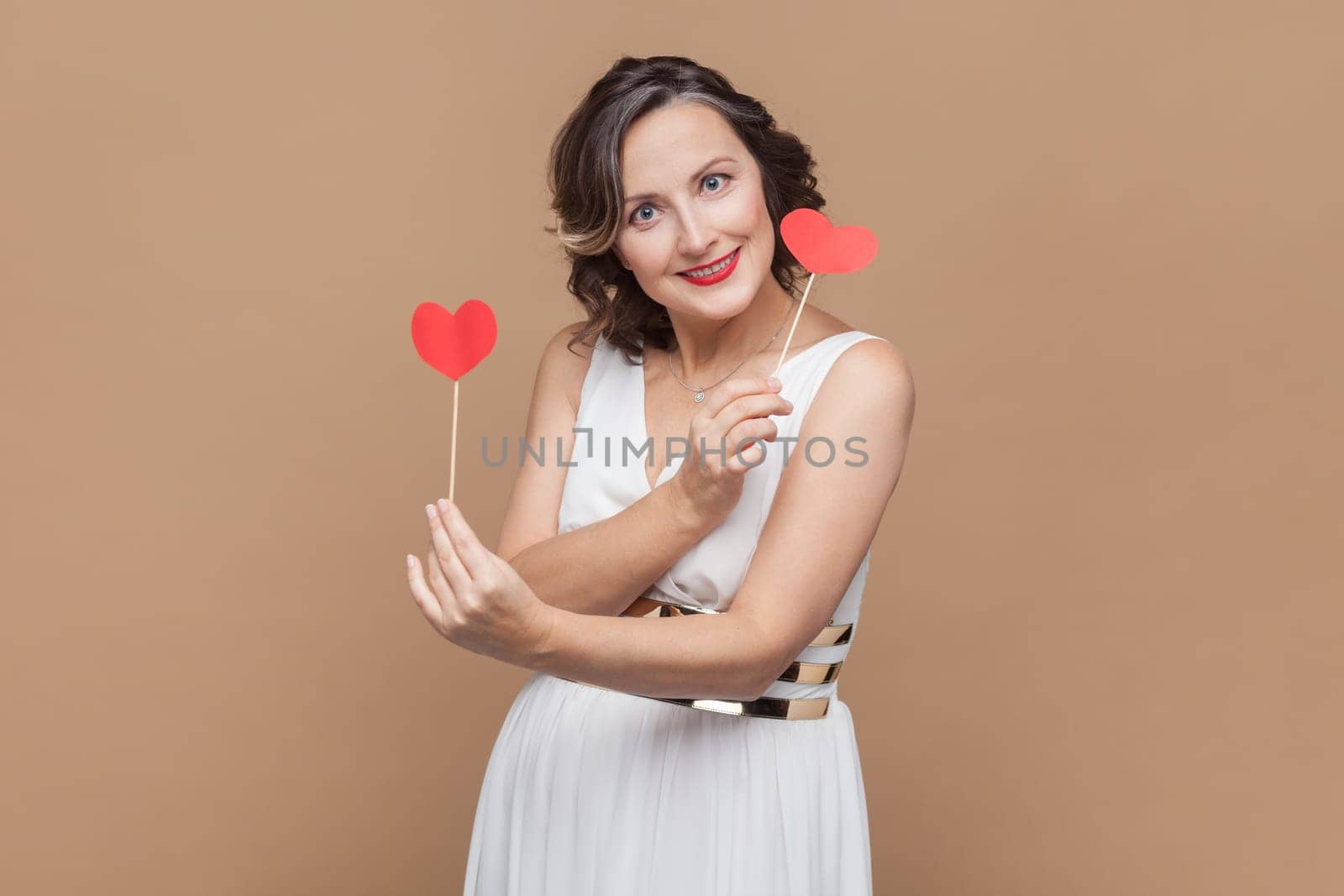 Portrait of cute charming middle aged woman with wavy hair holding little red hearts on sticks, looking smiling at camera, wearing white dress. Indoor studio shot isolated on light brown background.