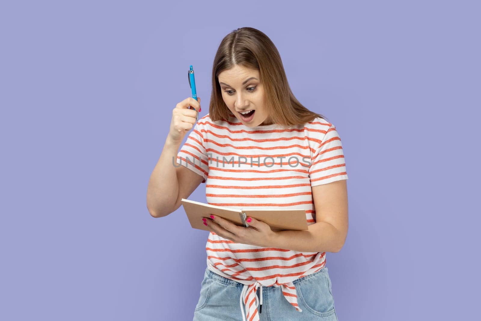 Portrait of excited amazed optimistic blond woman wearing striped T-shirt writing in paper notebook, raised pen, having excellent idea. Indoor studio shot isolated on purple background.