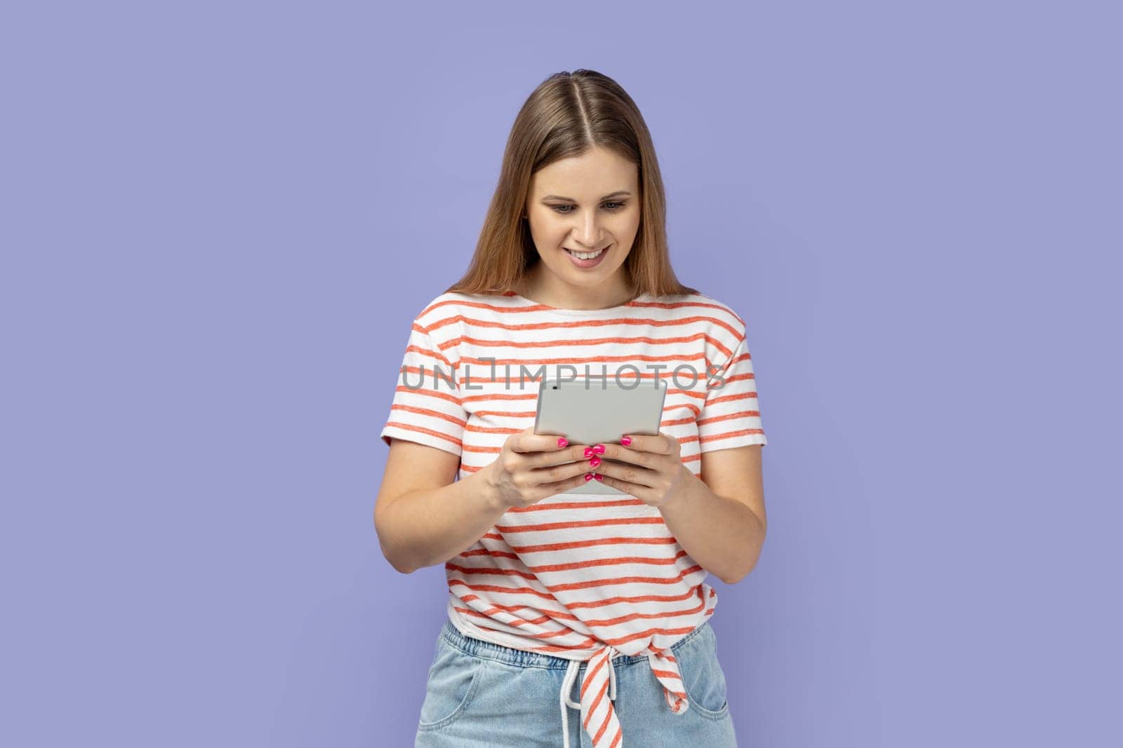 Portrait of adorable optimistic satisfied woman wearing striped T-shirt holding tablet, reading news or electronic book, looking at display with smile. Indoor studio shot isolated on purple background