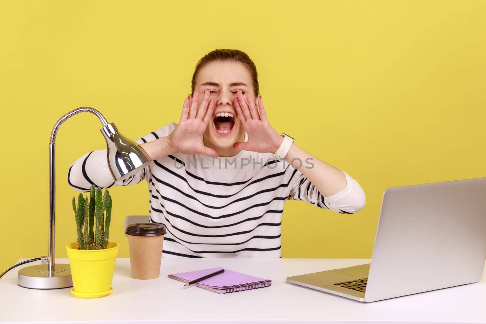 Nervous angry woman office worker in striped shirt loudly screaming holding hands near mouth, looking at camera, sitting at workplace. Indoor studio studio shot isolated on yellow background.