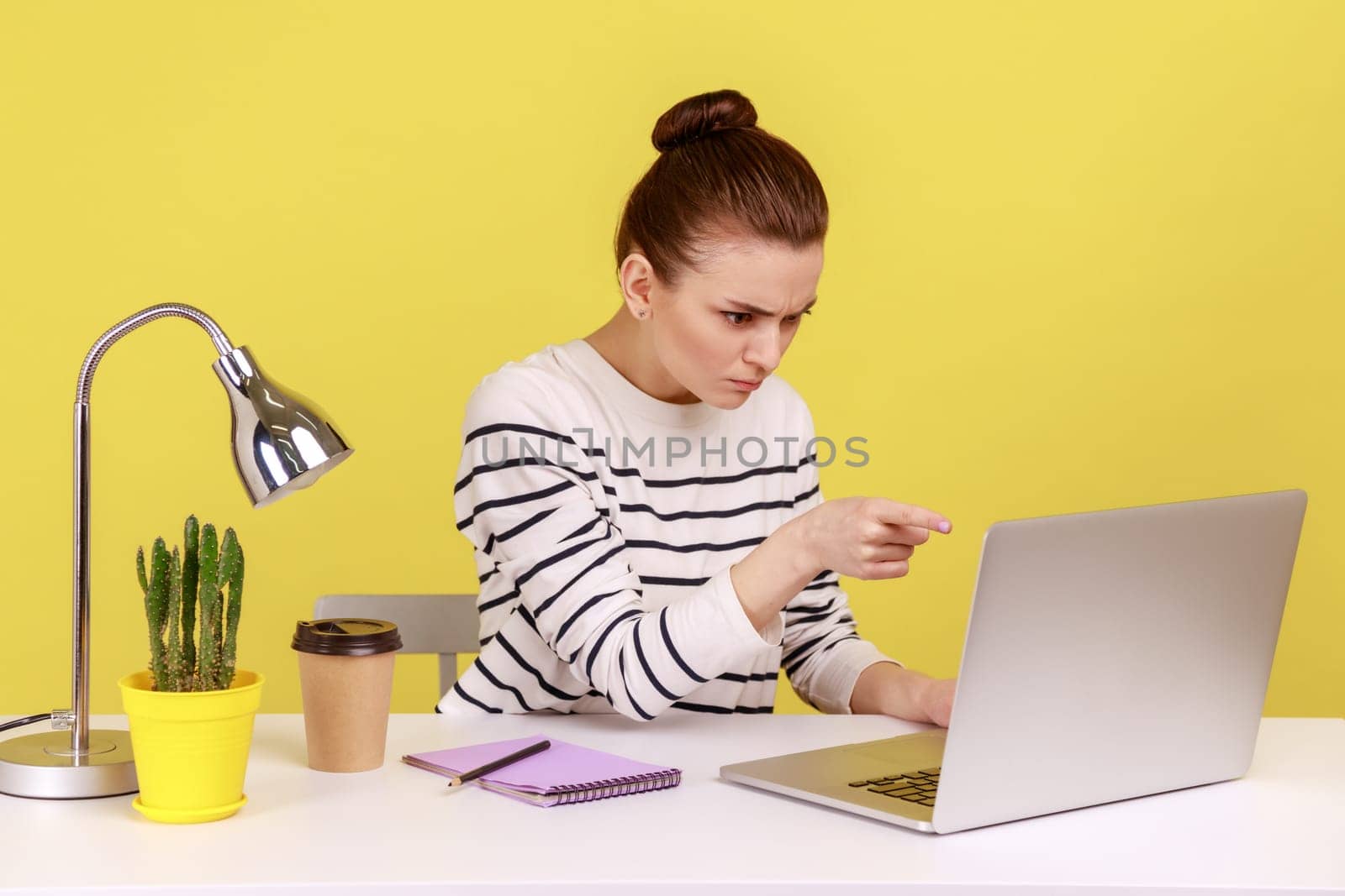 Angry woman office worker in striped shirt pointing to laptop screen, scolding and accusing talking on video call, having online conference. Indoor studio studio shot isolated on yellow background.