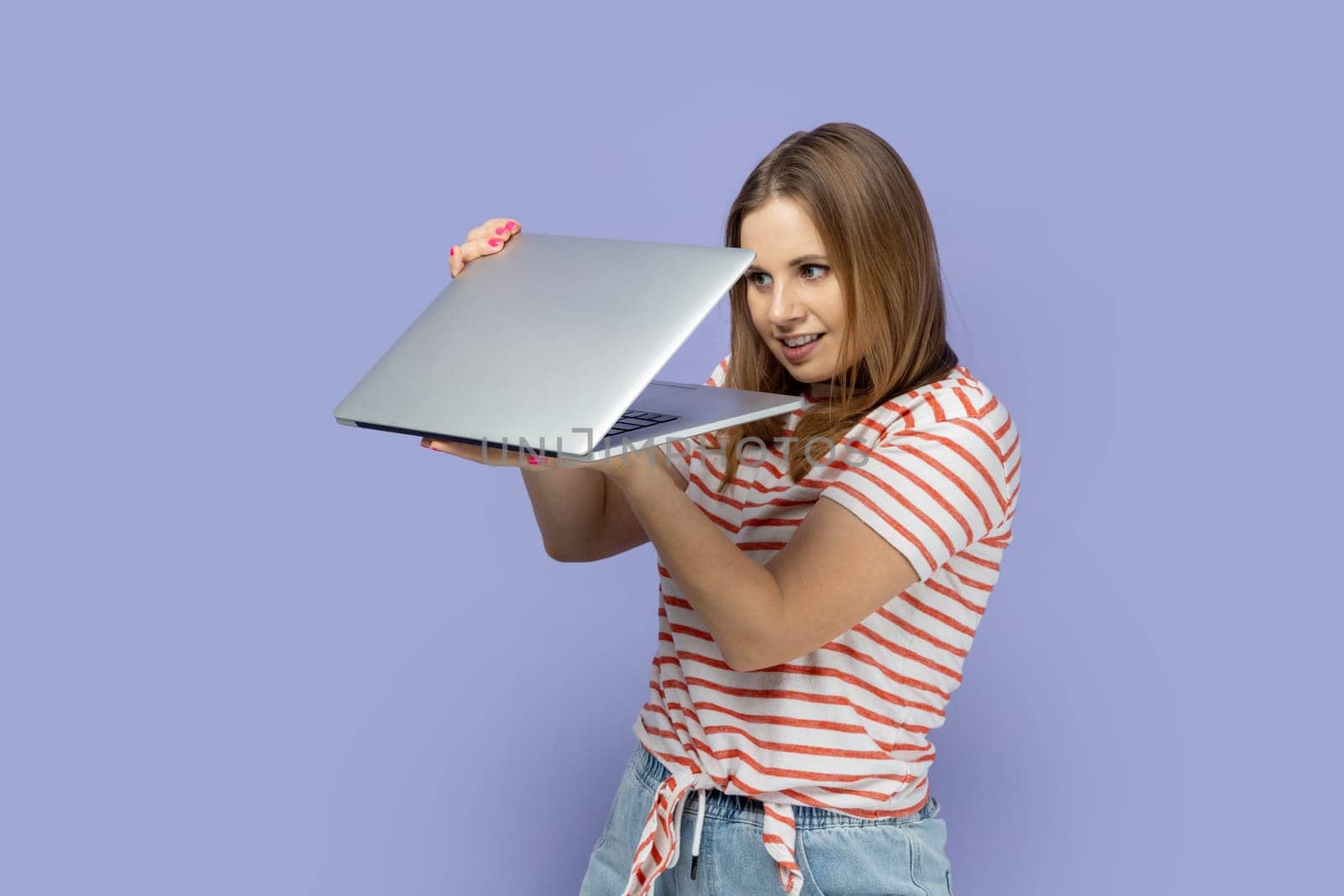 Portrait of curious blond woman wearing striped T-shirt holding half opened laptop, spying , finding important or forbidden information. Indoor studio shot isolated on purple background.