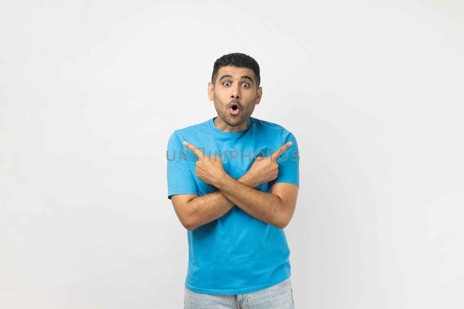 Portrait of shocked surprised astonished young adult unshaven man wearing blue T- shirt standing pointing with index fingers to different size. Indoor studio shot isolated on gray background.