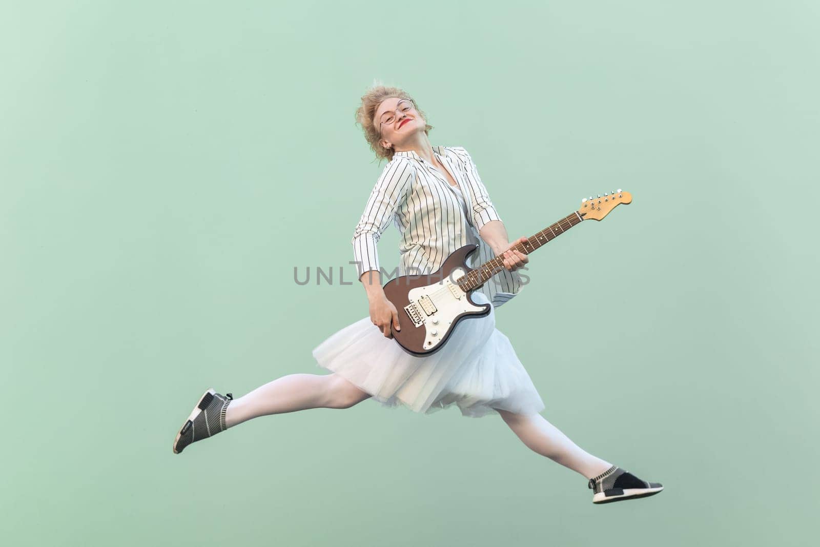 Happy blonde woman with eyeglasses holding electric guitar, smiling and jumping. by Khosro1