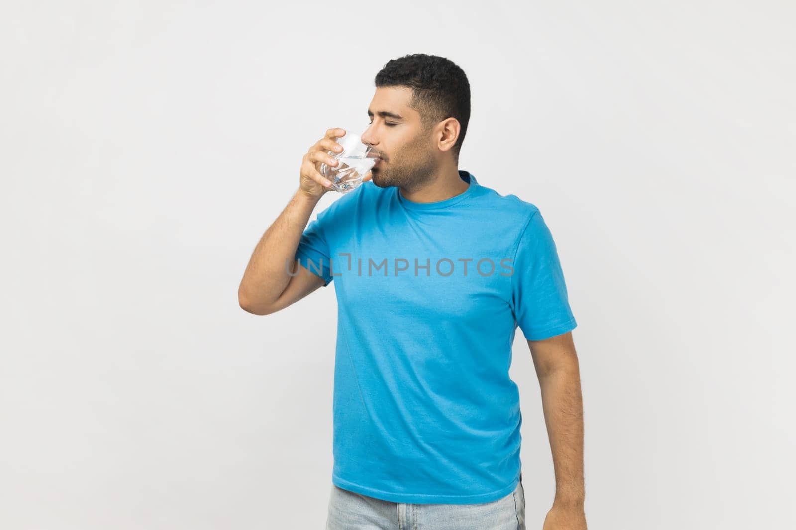 Portrait of thirsty handsome unshaven man in blue T- shirt standing with glass in hands, drinking fresh clean cold water during hot weather, enjoying. Indoor studio shot isolated on gray background.