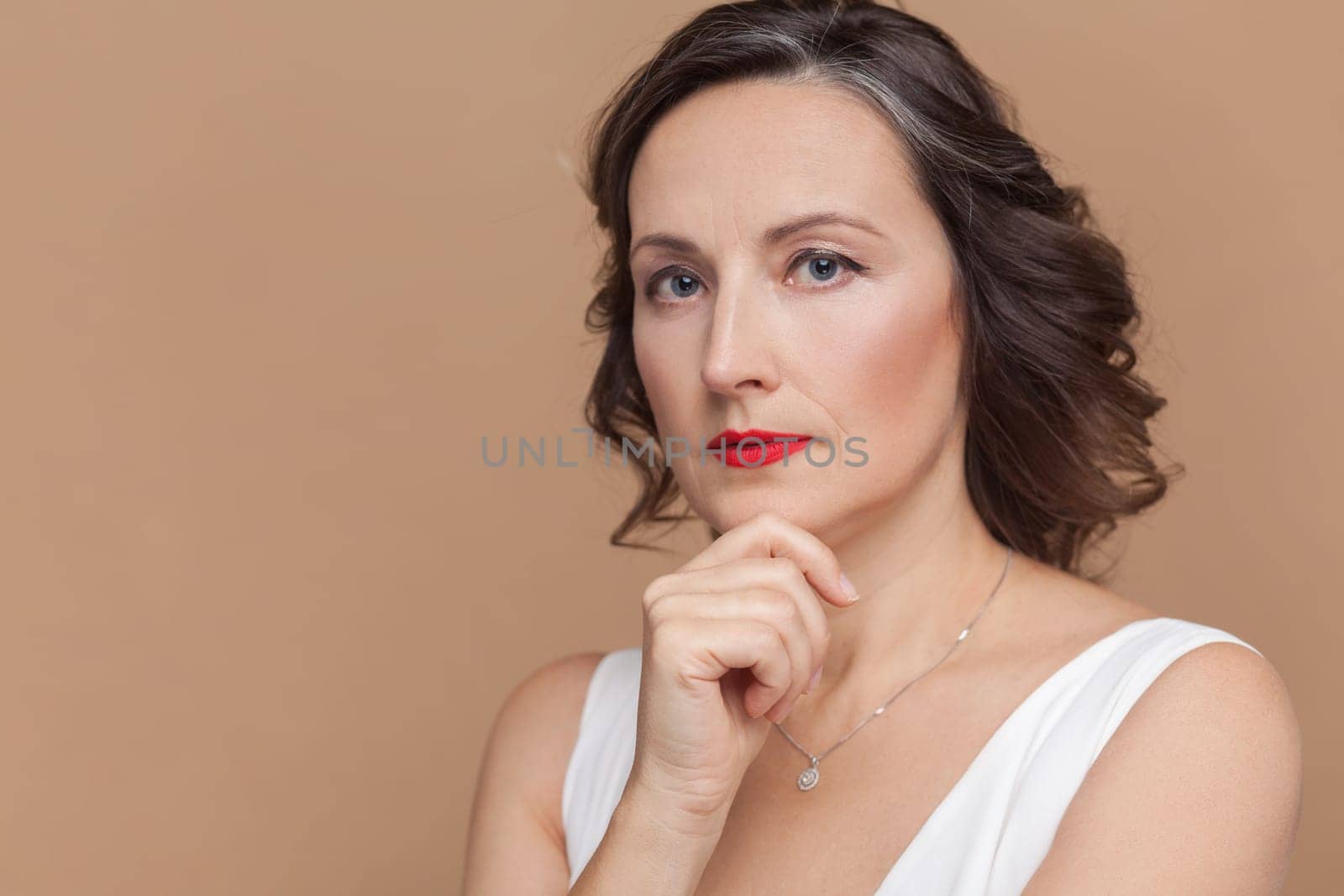 Closeup of serious pensive attractive beautiful middle aged woman with wavy hair and makeup, looking at camera, holding chin, wearing white dress. Indoor studio shot isolated on light brown background