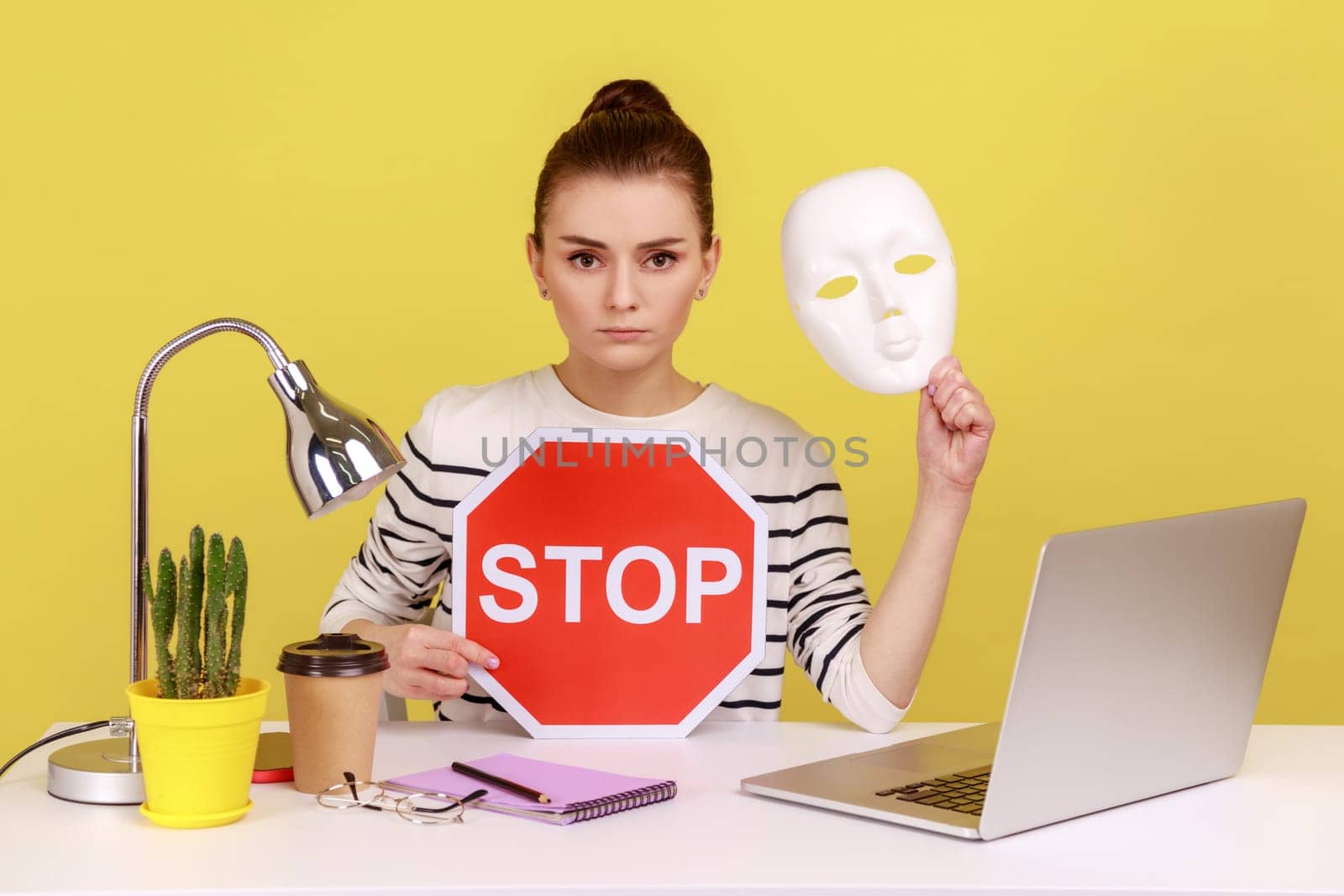 Portrait of serious dark haired woman holding white mask with unknown face and red traffic sign while sitting on workplace with laptop. Indoor studio studio shot isolated on yellow background.