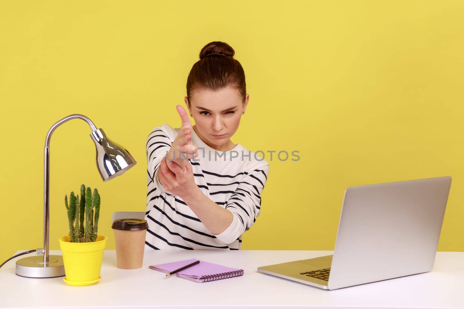 Angry woman office worker sitting at workplace with laptop threatening and pointing finger gun to camera, shooting with aggressive expression. Indoor studio studio shot isolated on yellow background.