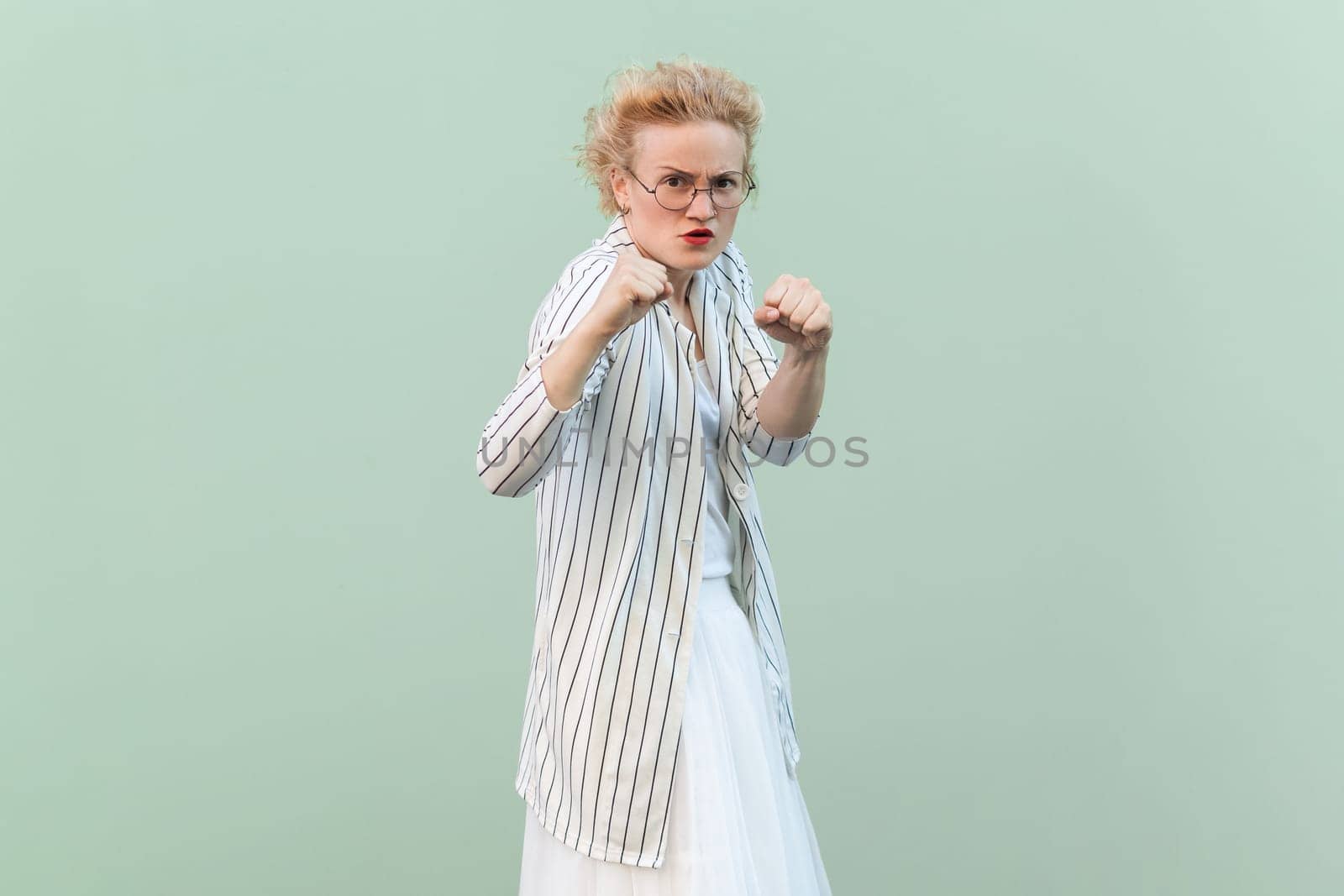 Portrait of angry aggressive attractive blonde woman wearing striped shirt and skirt, clenching fists, being ready to attack. Indoor studio shot isolated on light green background.