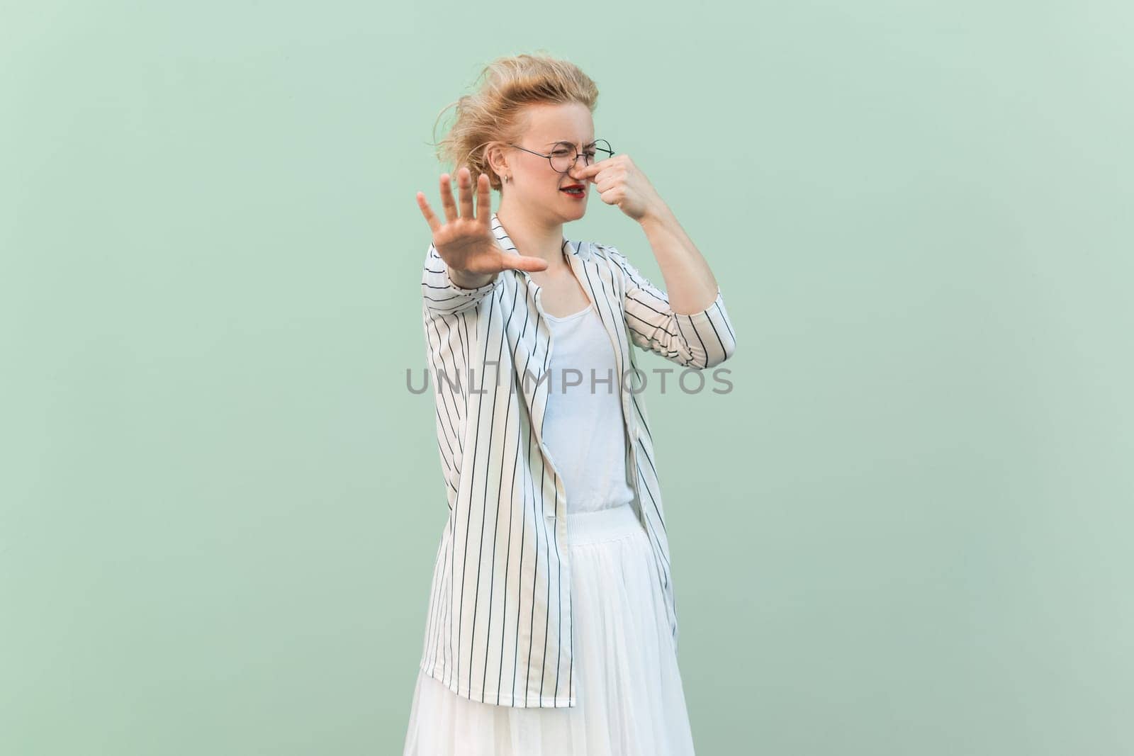 Portrait of confused disgusted blonde woman wearing striped shirt and skirt pinching her nose, showing stop gesture. Indoor studio shot isolated on light green background.