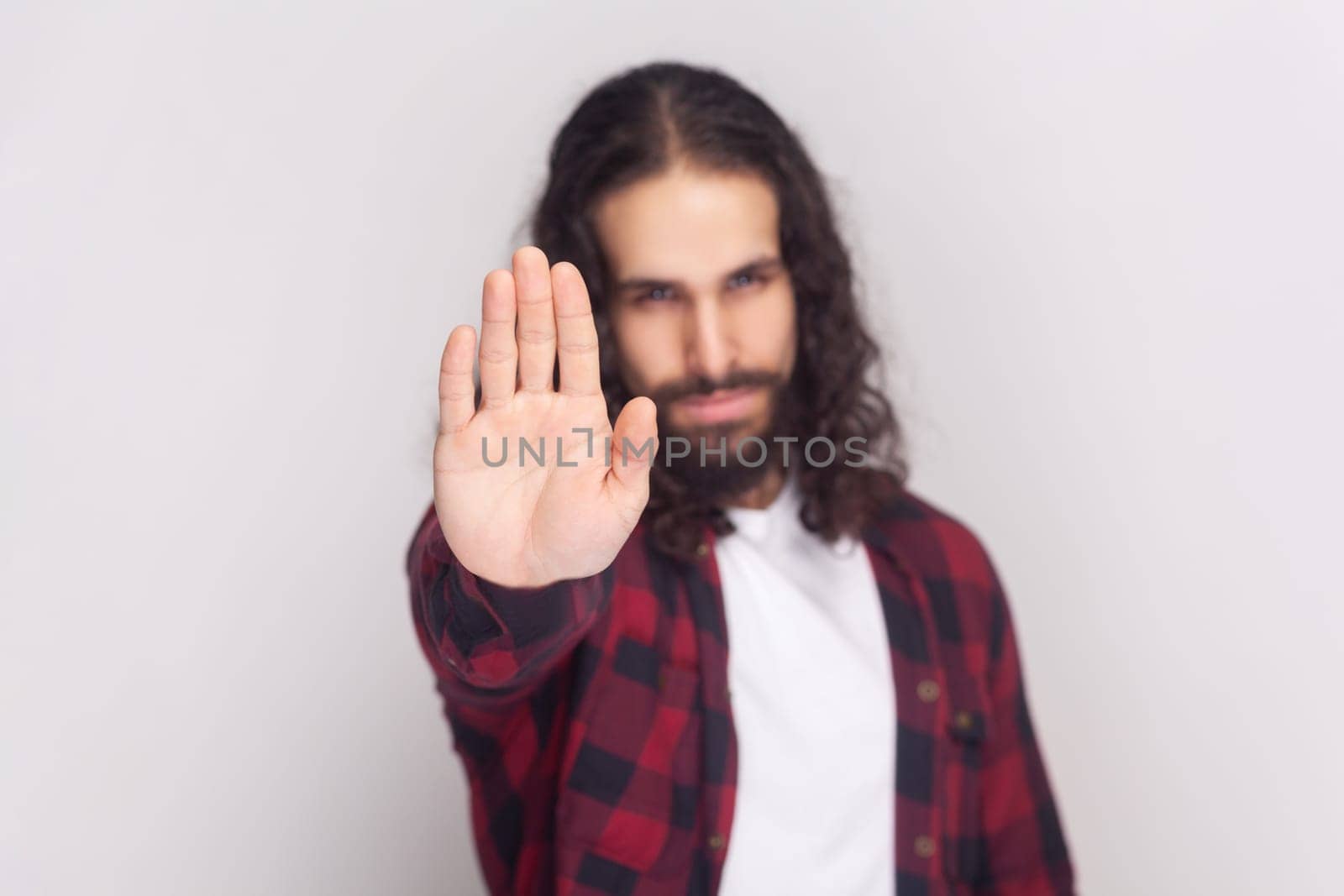 Serious bearded man with long curly hair in checkered red shirt does stop sign with palm, demonstrates prohibition and restriction, refuses something. Indoor studio shot isolated on gray background.