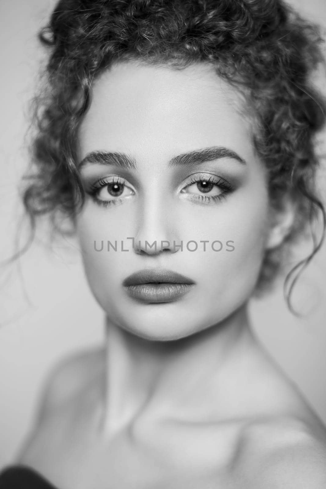 Black and white beauty portrait of young attractive woman fashion model with collected dark curly hair, looking at camera with serious face. Indoor studio shot isolated on gray background.