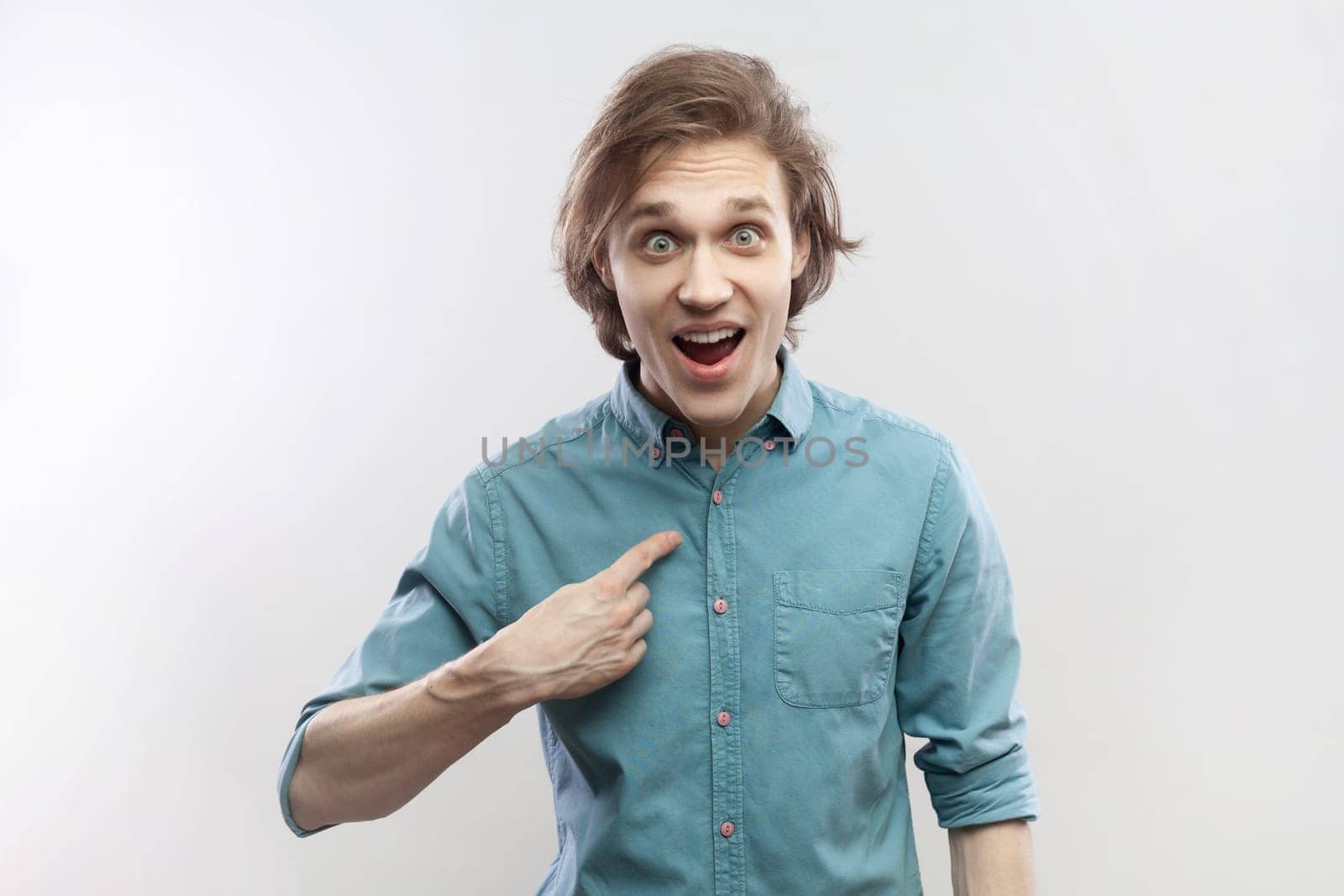 Portrait of young man pointing himself and looking at camera with surprised expression, shocked by her job achievement, wearing blue shirt. Indoor studio shot isolated on gray background.