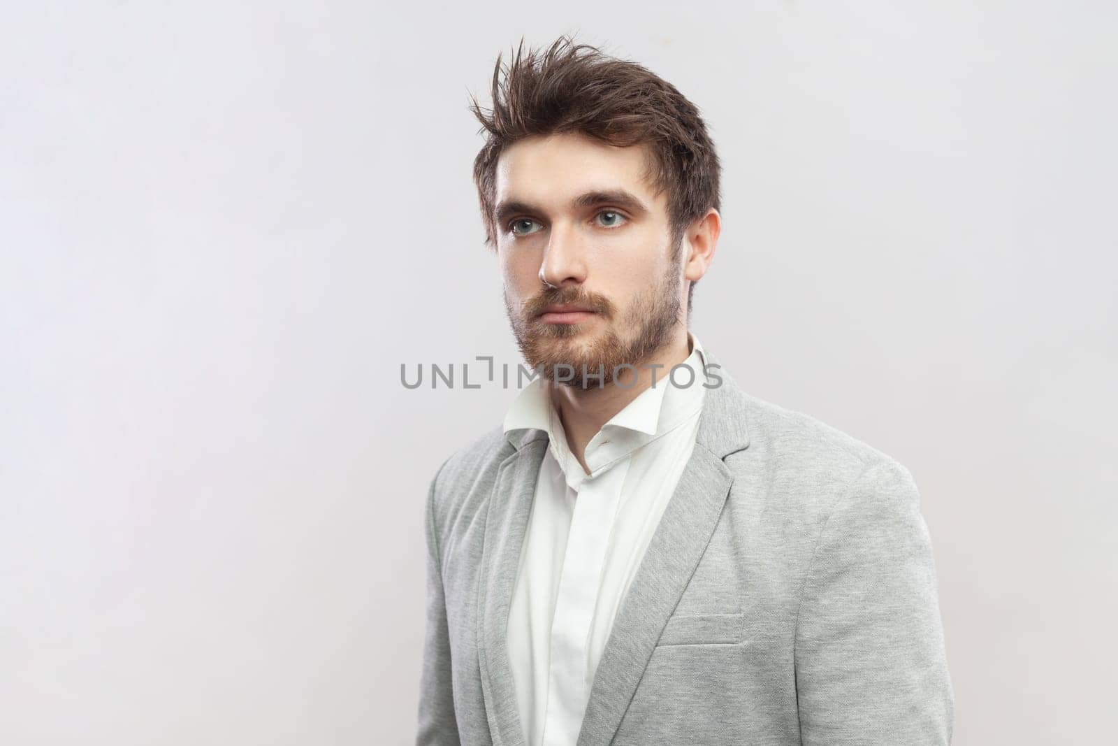 Portrait of handsome attractive young bearded man standing with serious concentrated facial expression, looking away, wearing grey suit. Indoor studio shot isolated on gray background.
