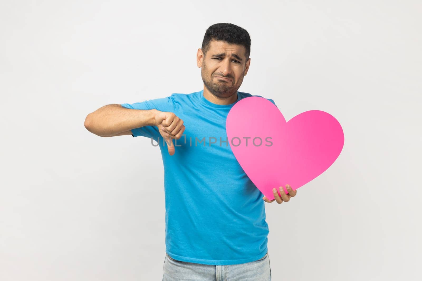Portrait of sad upset romantic unshaven man wearing blue T- shirt standing holding big pink heart and showing thumb down, dislike gesture. Indoor studio shot isolated on gray background.