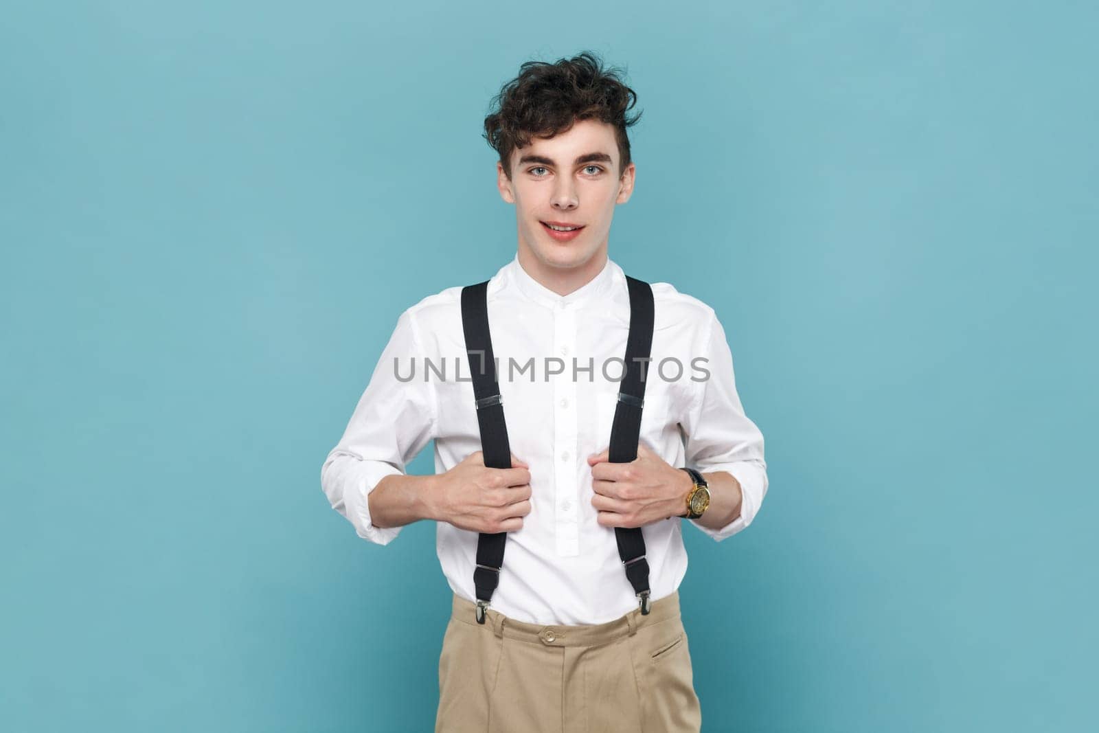 Portrait of smiling satisfied young man wearing white shirt holding his suspenders, looking at camera with positive facial expression. Indoor studio shot isolated on blue background.