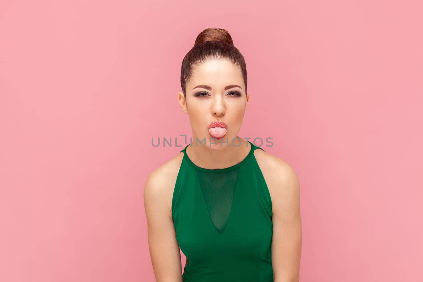 Portrait of funny attractive woman with bun hairstyle, showing tongue out, demonstrating childish behavior, wearing green dress. Indoor studio shot isolated on pink background.