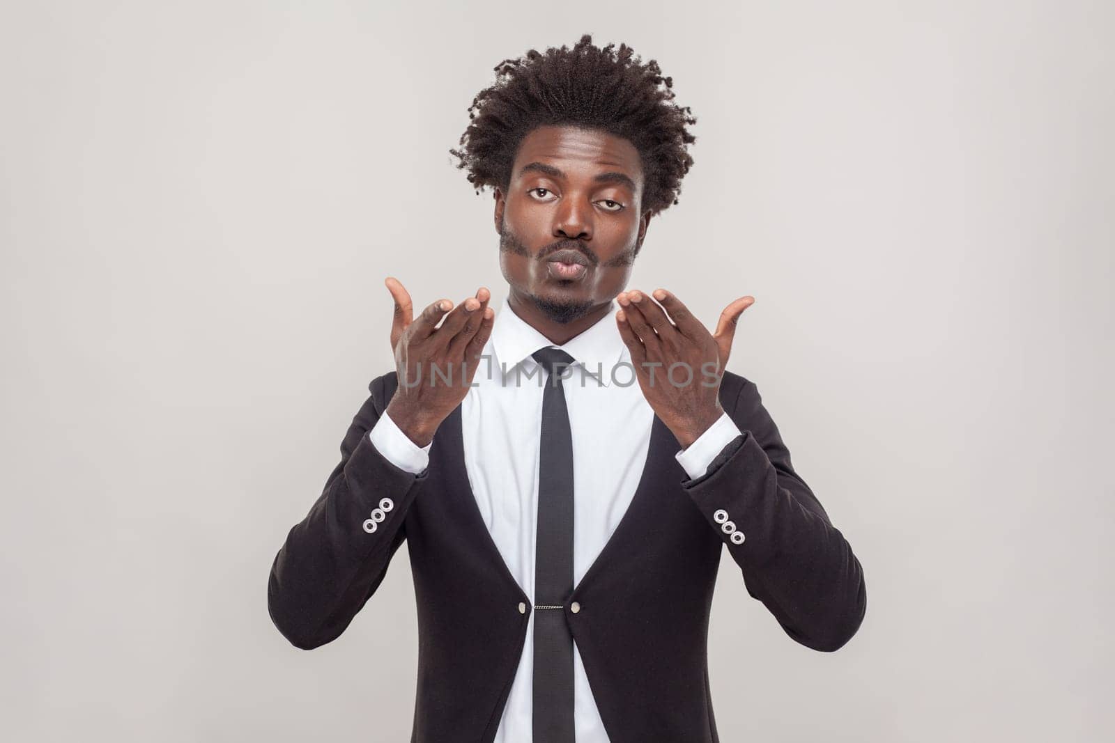 Portrait of pleased man with Afro hairstyle stretches hands near face, blows air kiss, expresses love and affection, wearing white shirt and tuxedo. Indoor studio shot isolated on gray background.