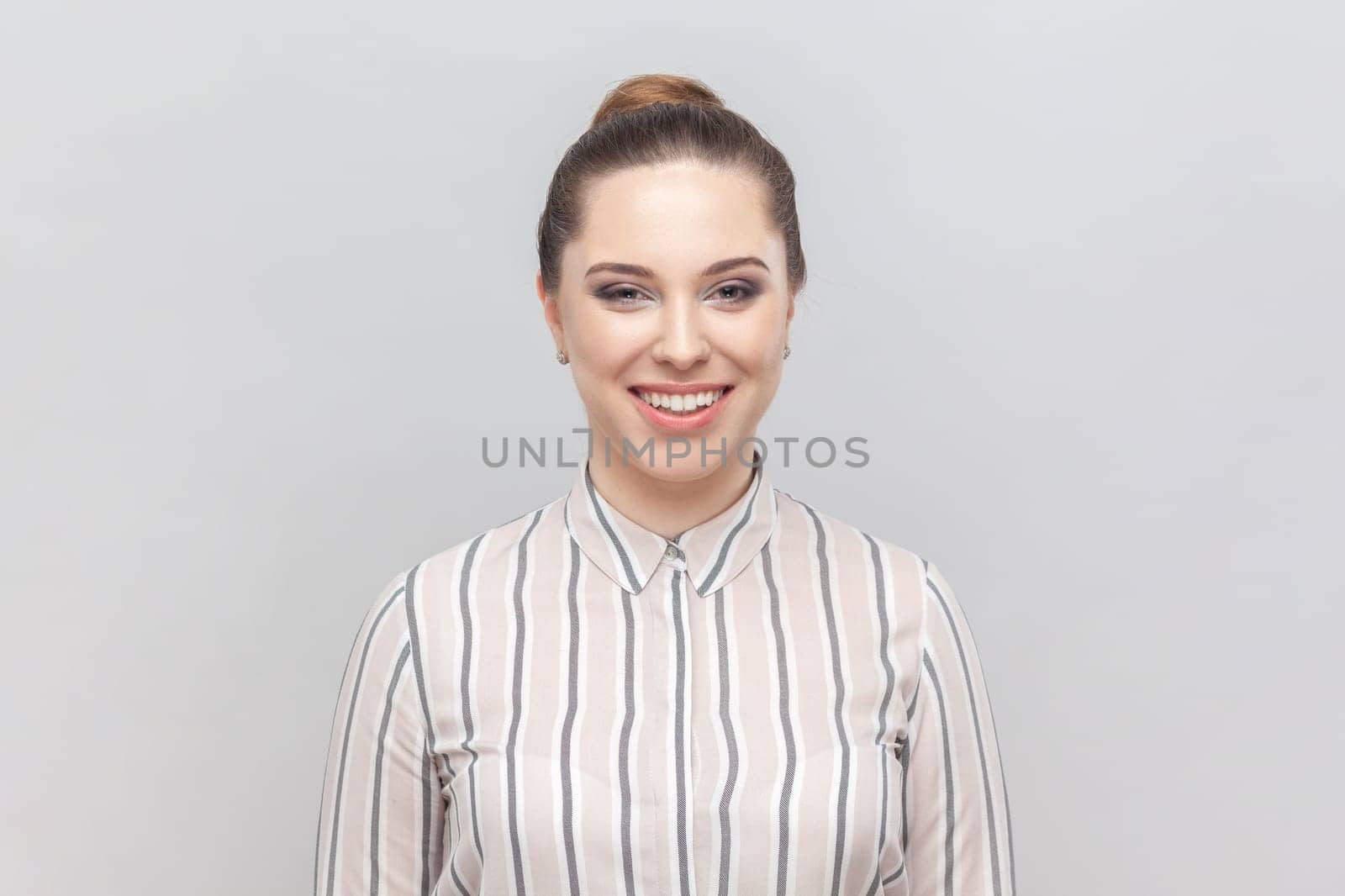 Portrait of attractive joyful cheerful woman wearing striped shirt looking at camera with toothy smile, being in good mood, expressing positiveness. Indoor studio shot isolated on gray background.