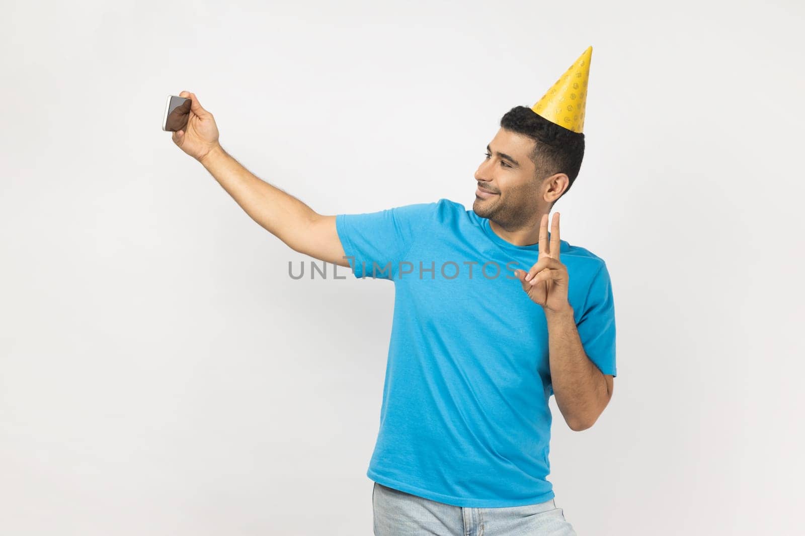 Portrait of friendly unshaven man blogger wearing blue T- shirt and yellow party cone making selfie or broadcasting livestream, showing v sign. Indoor studio shot isolated on gray background.