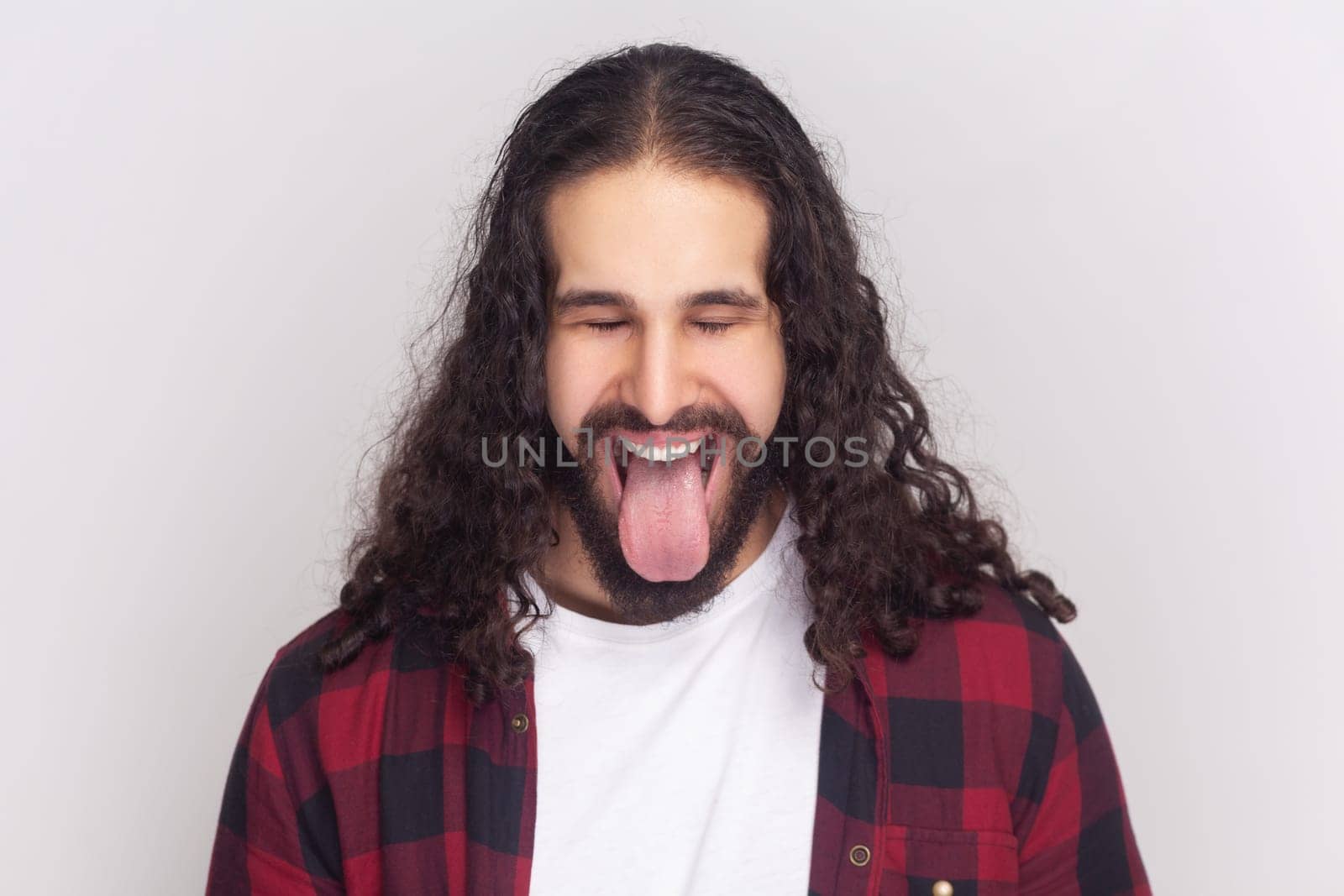 Comic bearded man in checkered red shirt sticks out tongue, makes happy grimace, expresses positive emotions, foolishes with friends. Indoor studio shot isolated on gray background.