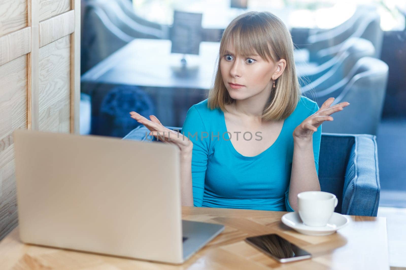 Portrait of unhappy serious attractive young woman with blonde hair in blue shirt working on laptop, having video call, spreads hands, asking how could you. Indoor shot, cafe background.