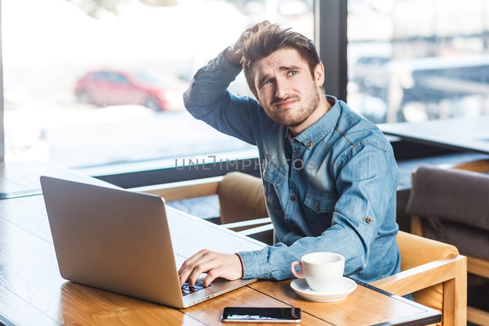 Portrait of pensive attractive young man freelancer in blue jeans shirt working on laptop, thinking about idea for presentation. Indoor shot near big window, cafe background.