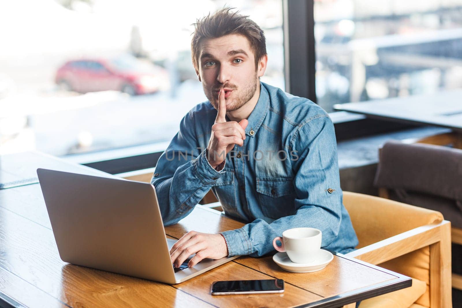 Portrait of handsome bearded young man freelancer in blue jeans shirt working on laptop, showing shh gesture, looking at camera, asking not to make noise. Indoor shot near big window, cafe background.