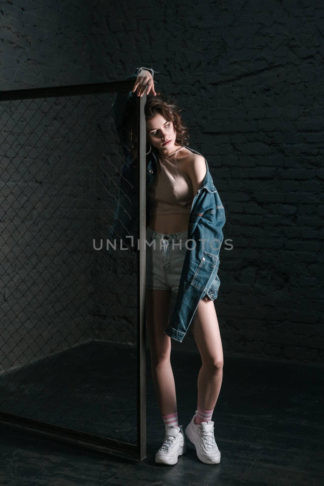 Full length portrait of beautiful pensive thoughtful woman wearing shorts and denim jacket, looking away standing leaning on fence on dark brick wall background. Indoor studio shot.