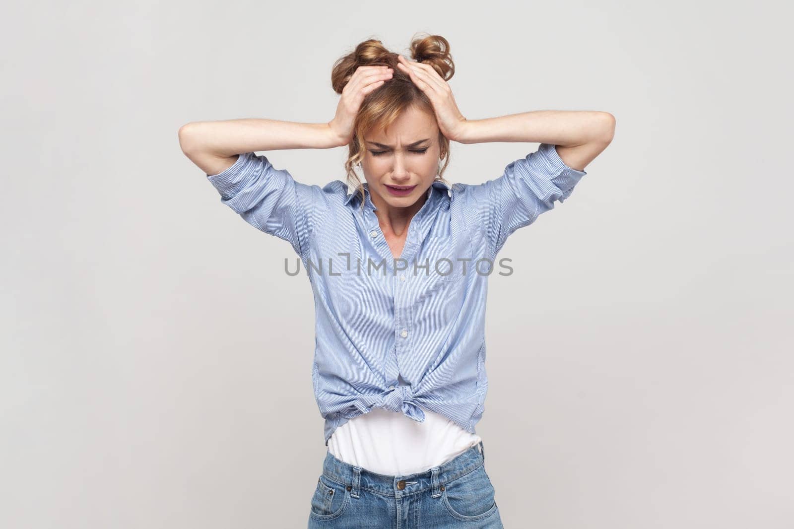 Portrait of blonde woman suffering from migraine, touching head, needs painkillers, cannot hold pain, feels discomfort, wearing blue shirt. Indoor studio shot isolated on gray background.