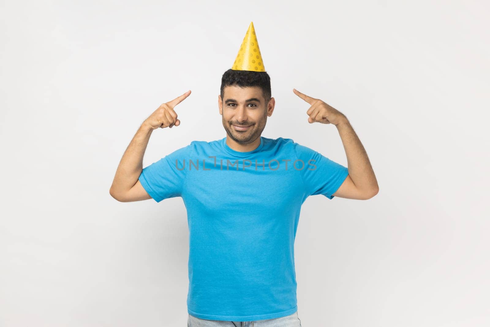 Portrait of festive positive optimistic unshaven man wearing blue T- shirt standing and pointing at yellow party cone, celebrating his birthday. Indoor studio shot isolated on gray background.