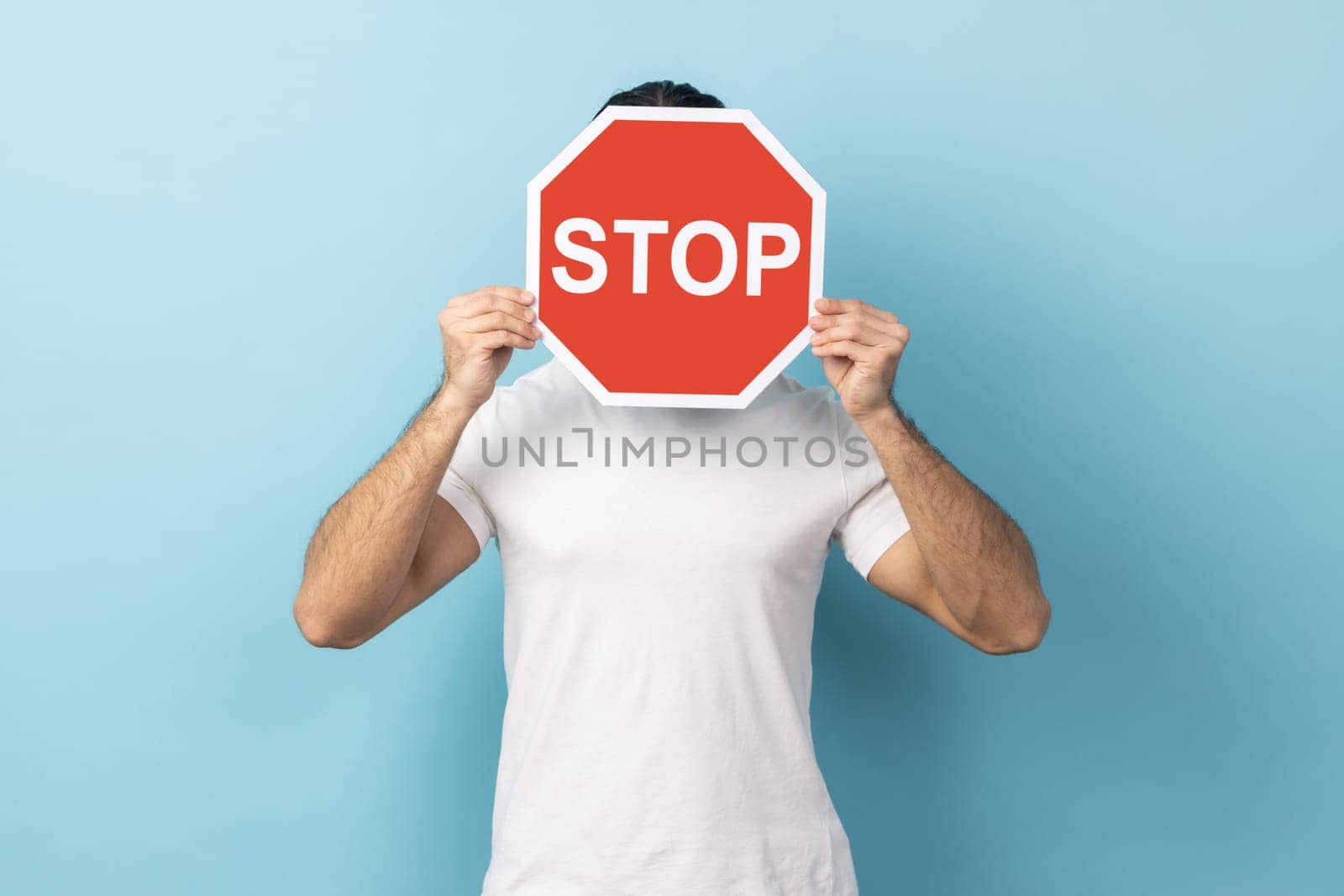 Portrait of unknown man wearing white T-shirt covering face with Stop symbol, anonymous person holding red traffic sign, warning about road safety rules. Indoor studio shot isolated on blue background