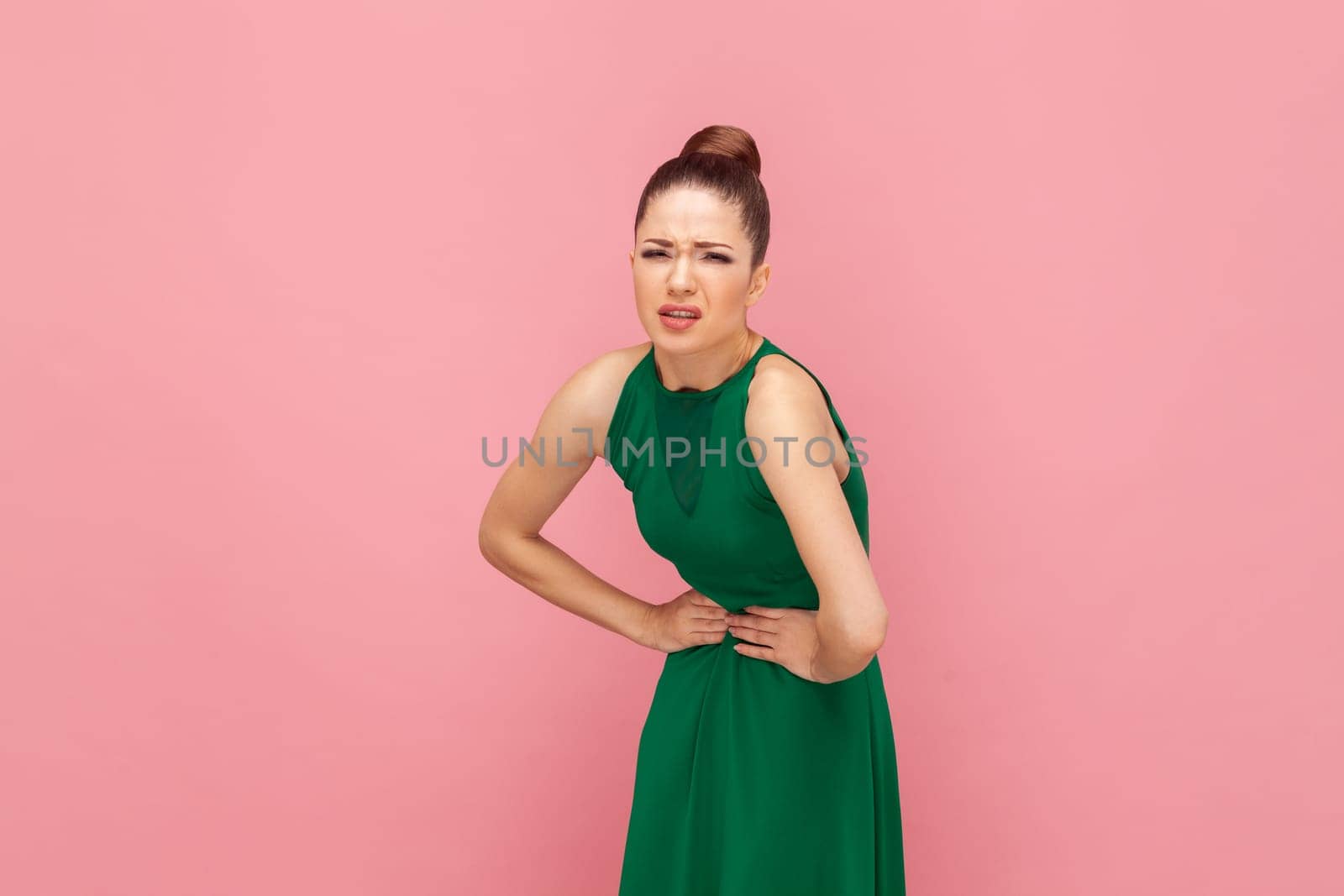 Portrait of dissatisfied unhealthy sick woman keeping hands on stomach, suffers from pain, has menstruation, wearing green dress. Indoor studio shot isolated on pink background.