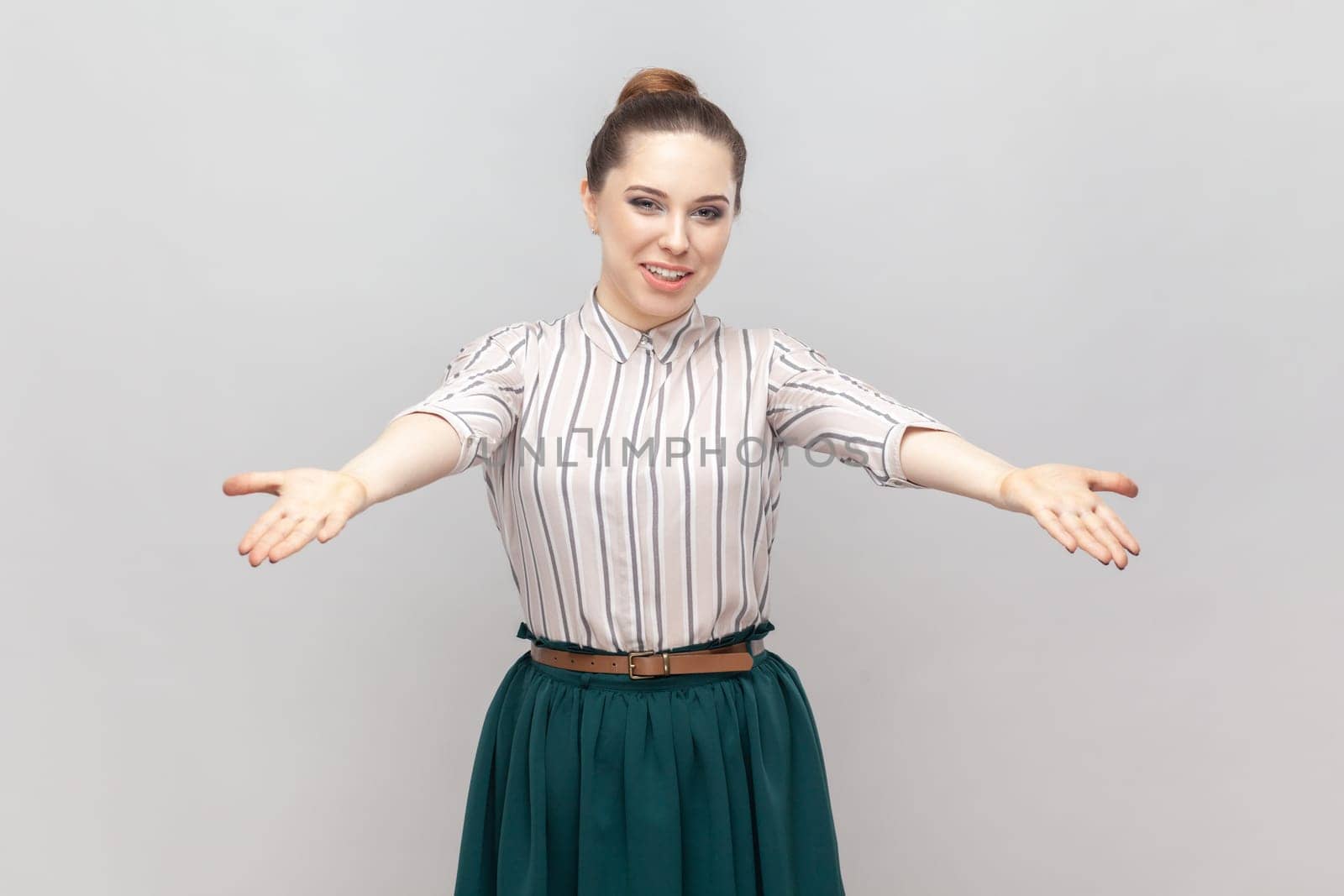 Please, take it for free. Portrait of pleased friendly optimistic woman wearing striped shirt and green skirt standing with spread hands. Indoor studio shot isolated on gray background.