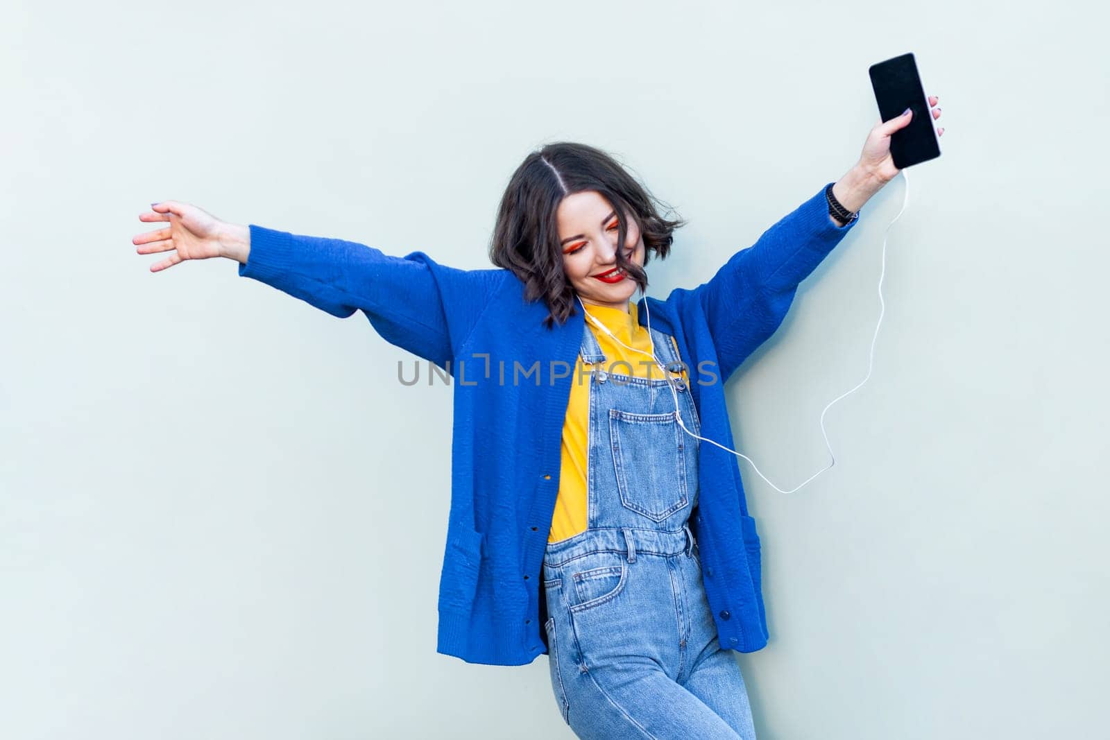 Portrait of joyful hipster woman in denim overalls yellow T-shirt and blue jacket listening to music on smart phone, keeps eyes closed, enjoying. Indoor studio shot isolated on light green background