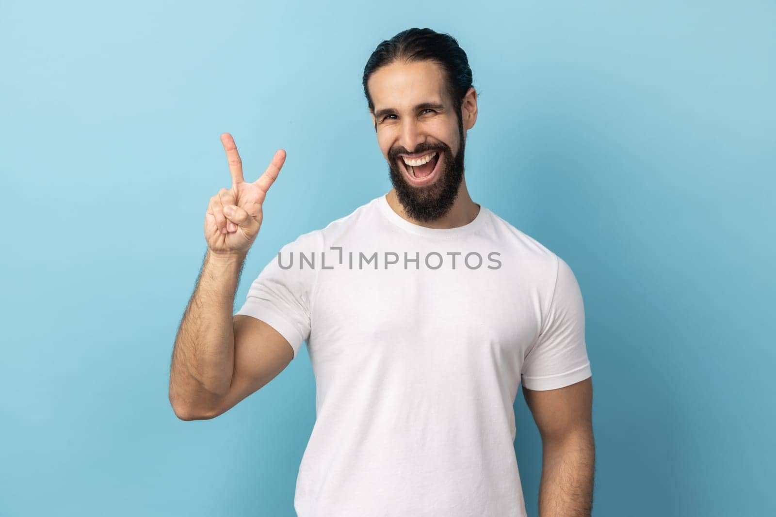 Portrait of positive man with beard wearing white T-shirt looking at camera and gesturing victory sign at camera, success or achievement. Indoor studio shot isolated on blue background.