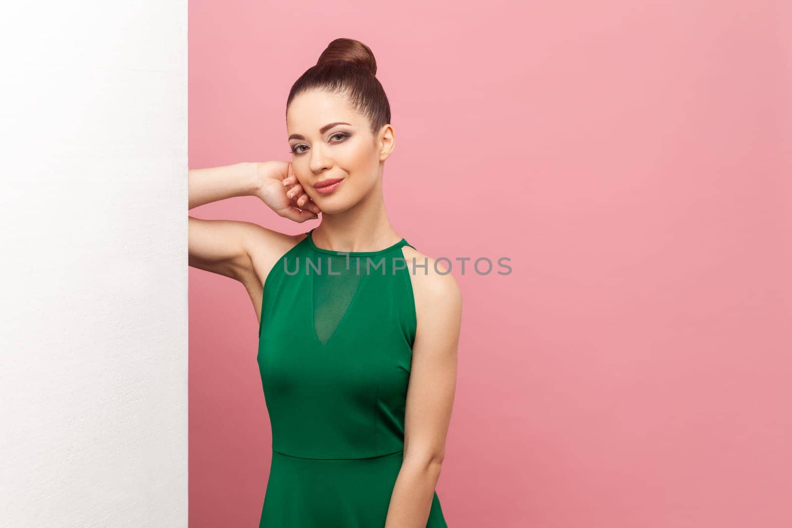 Portrait of attractive winsome beautiful young adult woman with bun hairstyle standing leaning on white wall keeps hand near face, wearing green dress. Indoor studio shot