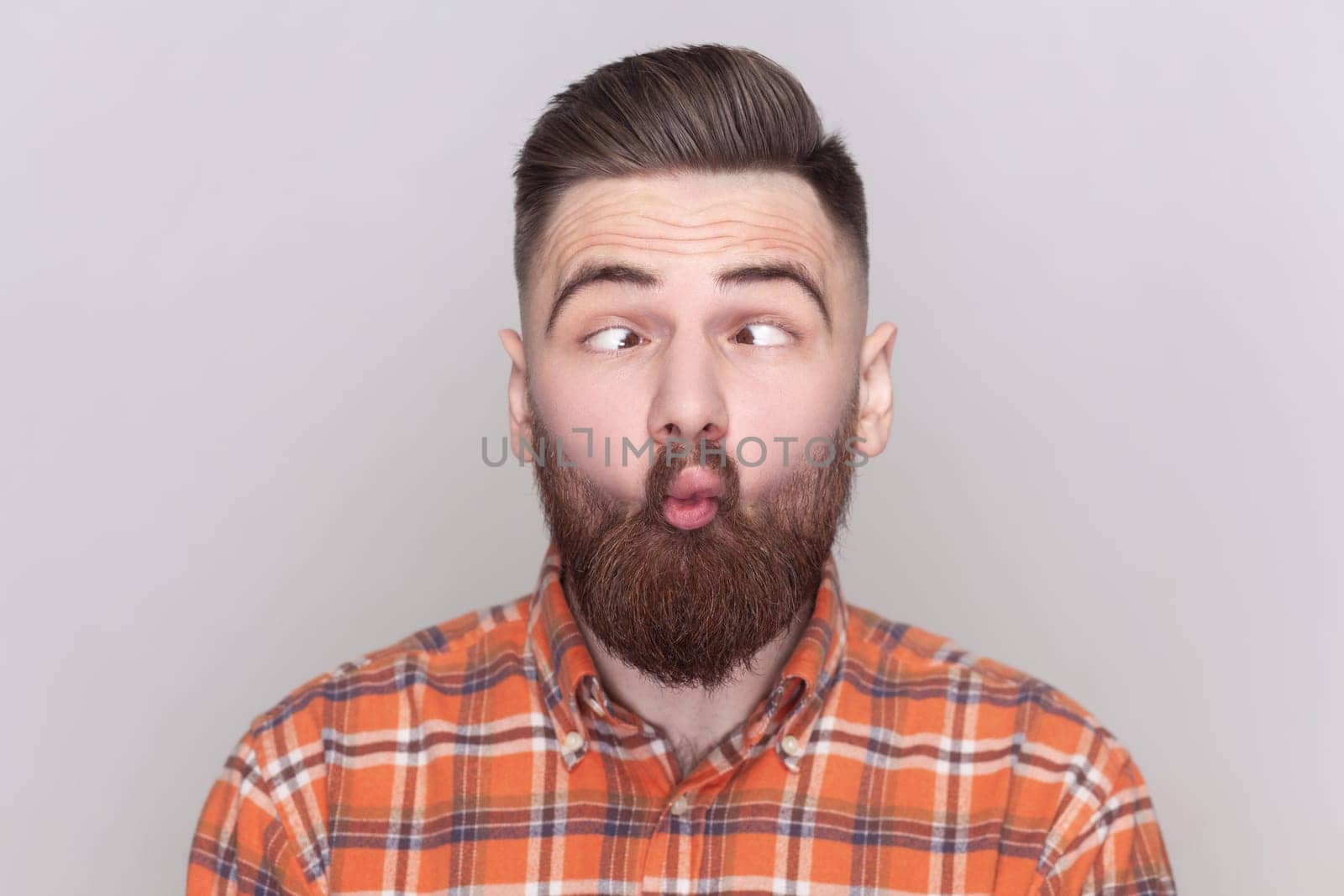 Portrait of funny crazy bearded man standing, crossed eyes with fish lips gesture, fool around, having fun, wearing checkered shirt. Indoor studio shot isolated on gray background.