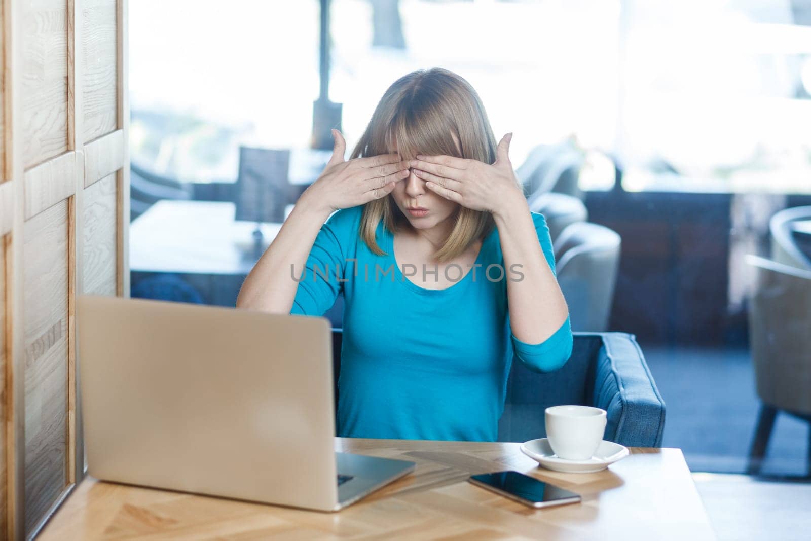 Portrait of attractive young woman with blonde hair in blue shirt working on laptop, covering eyes with palms, doesn't want to see something shameful. Indoor shot in cafe with big window on background