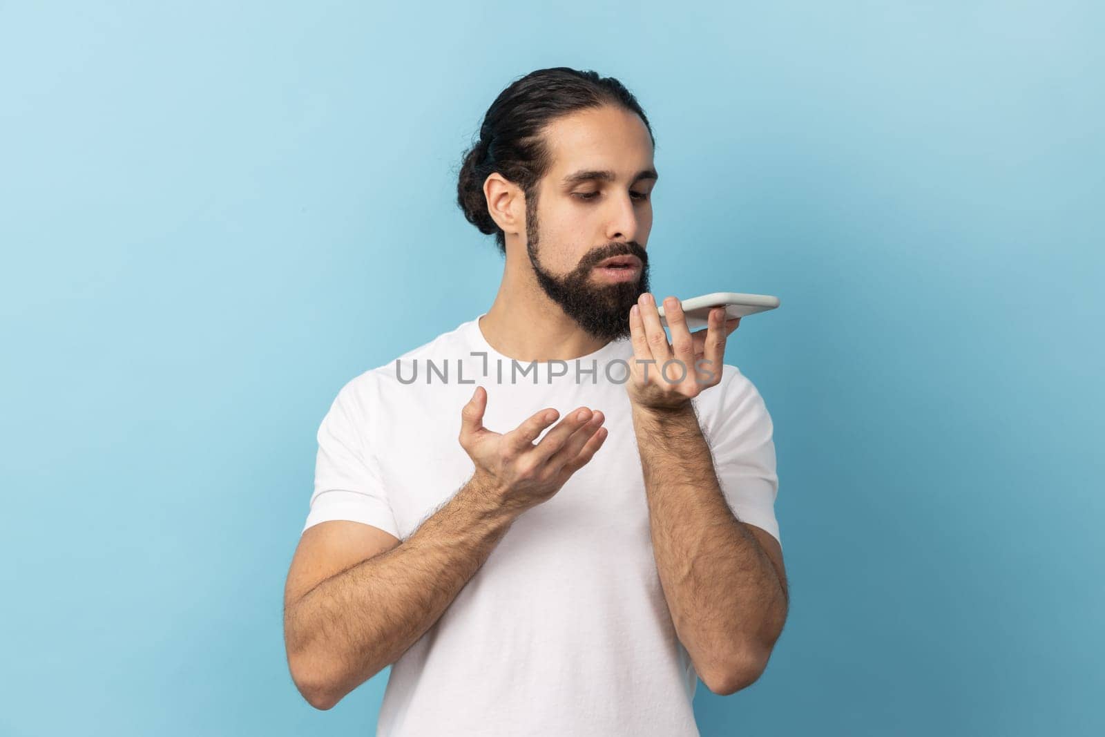 Smart voice technology. Portrait of man with beard wearing white T-shirt talking to cell phone using virtual assistant, digital speaker application. Indoor studio shot isolated on blue background.