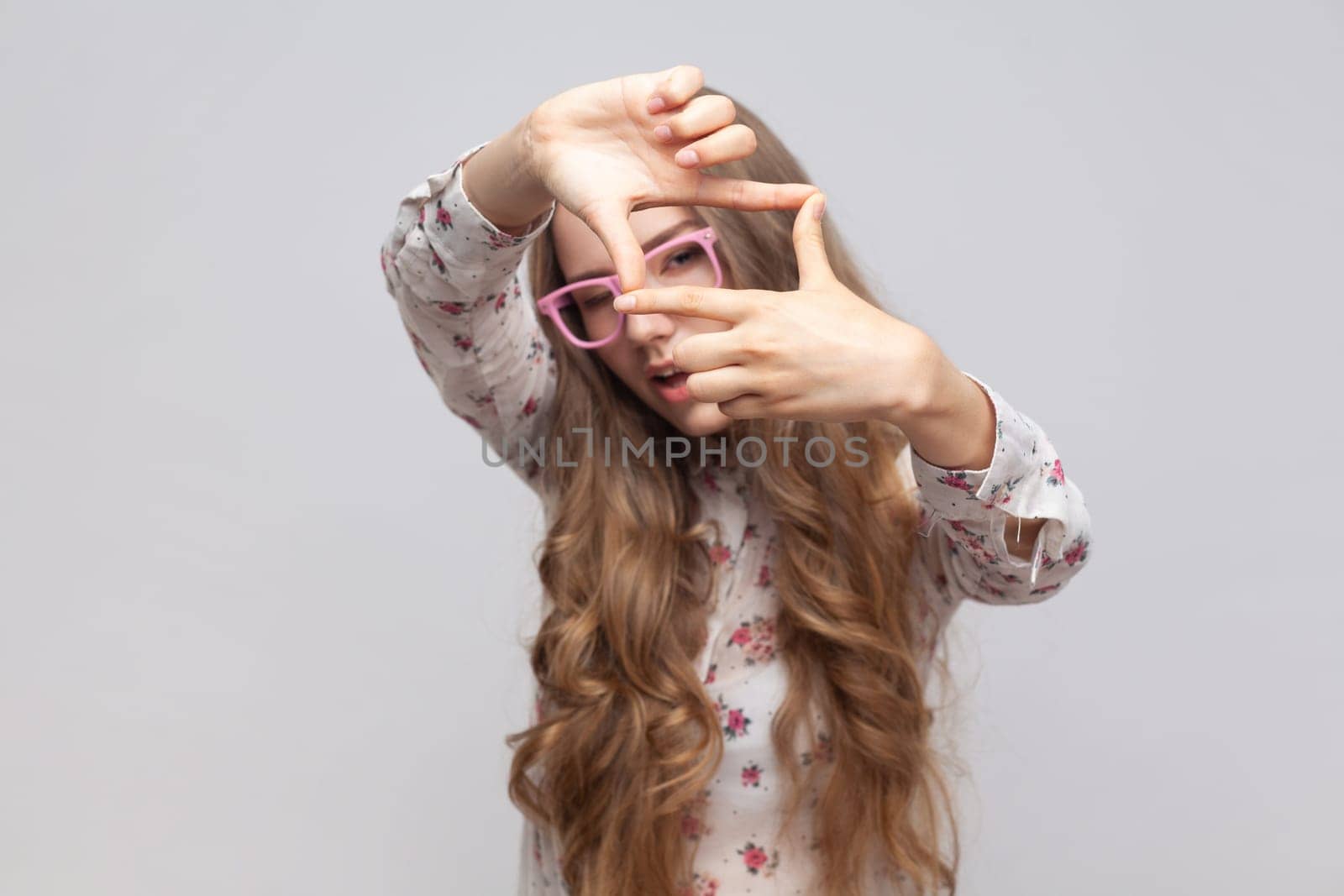 Woman in glasses with wavy blond hair gesturing picture frame with hands, looking through fingers and focusing on interesting moment, taking photo. Indoor studio shot isolated on gray background.