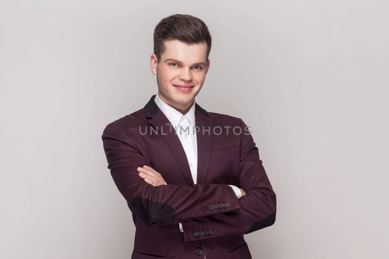 Portrait of confident clever handsome young man standing with crossed arms, looking at camera with happy expression, wearing violet suit and white shirt. Indoor studio shot isolated on grey background