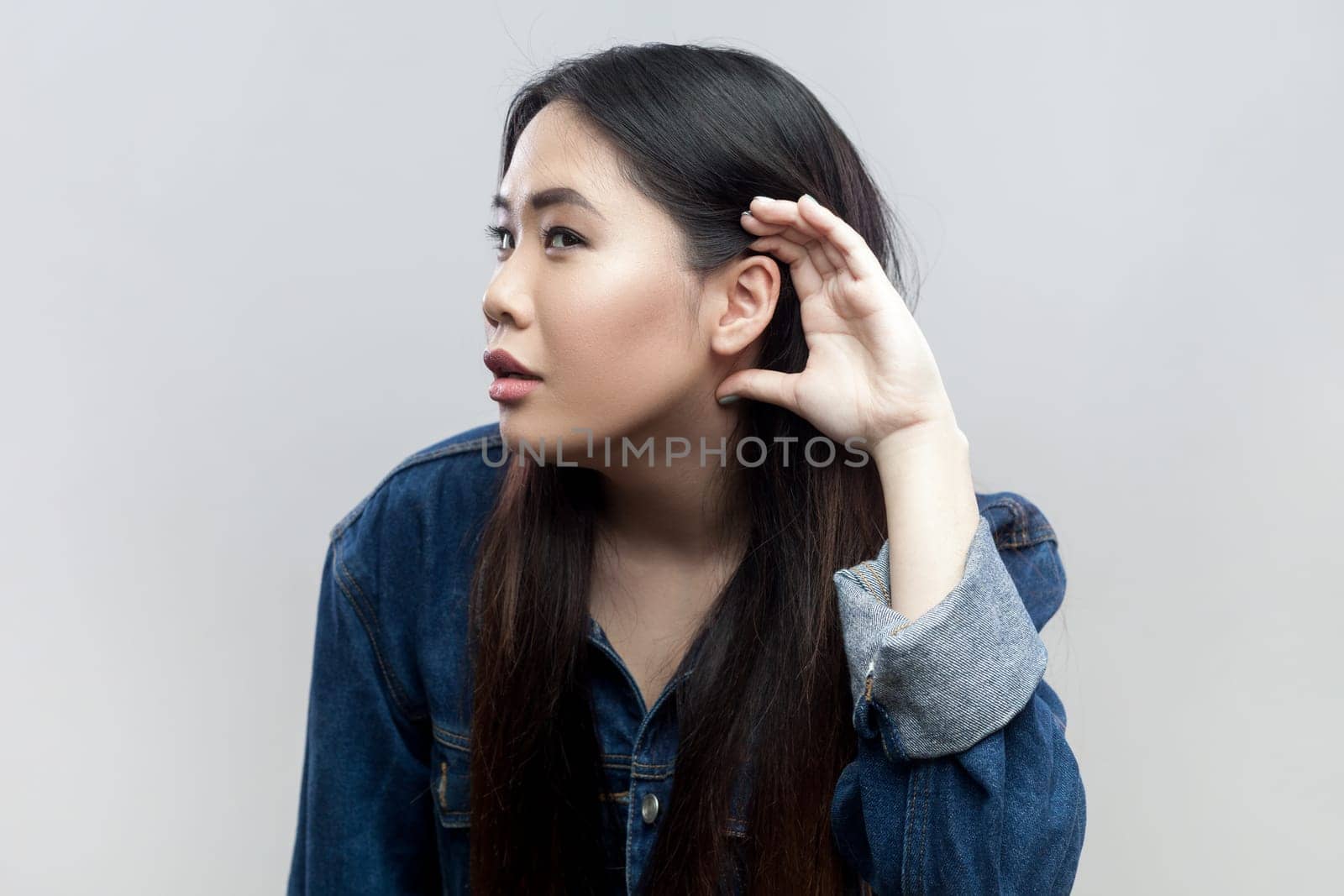 Portrait of brunette woman in blue denim jacket standing holding hand near ear, listening to interesting talks and private secrets, spying. Indoor studio shot isolated on gray background.