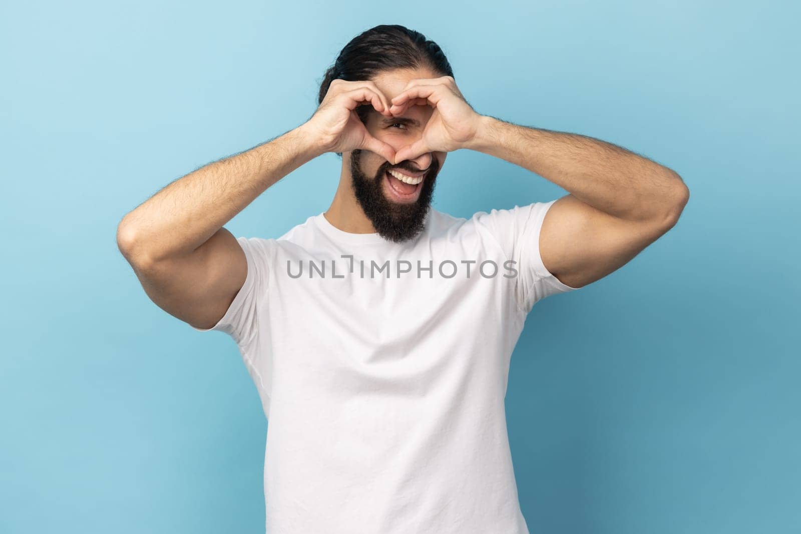 Portrait of happy pleased man with beard wearing white T-shirt making heart gesture over mouth smiles toothily, expressing romantic feelings. Indoor studio shot isolated on blue background.